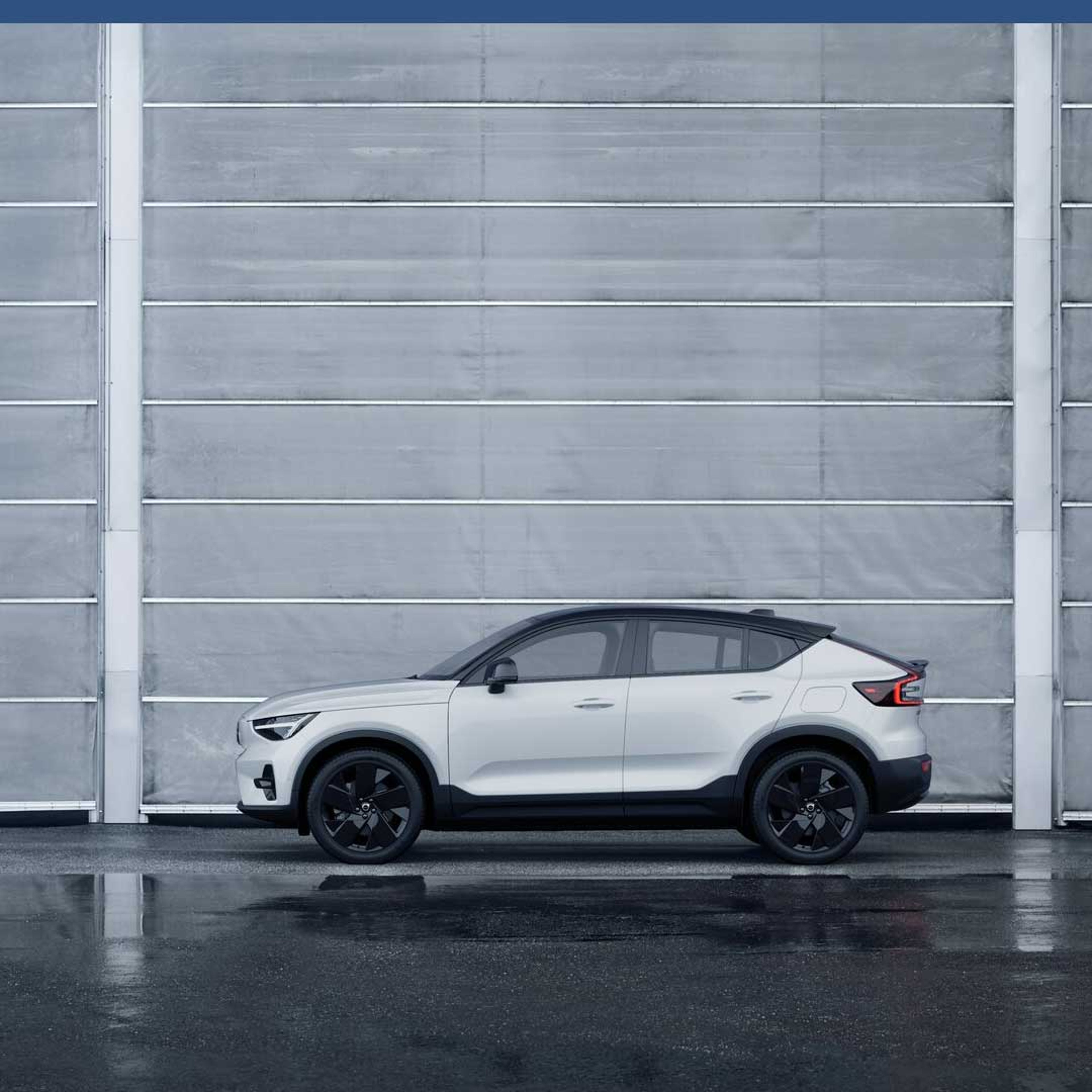 En elegant hvid Volvo SUV med sorte hjul står parkeret foran en stor grå industriport på våd asfalt og fremviser et moderne og minimalistisk design.