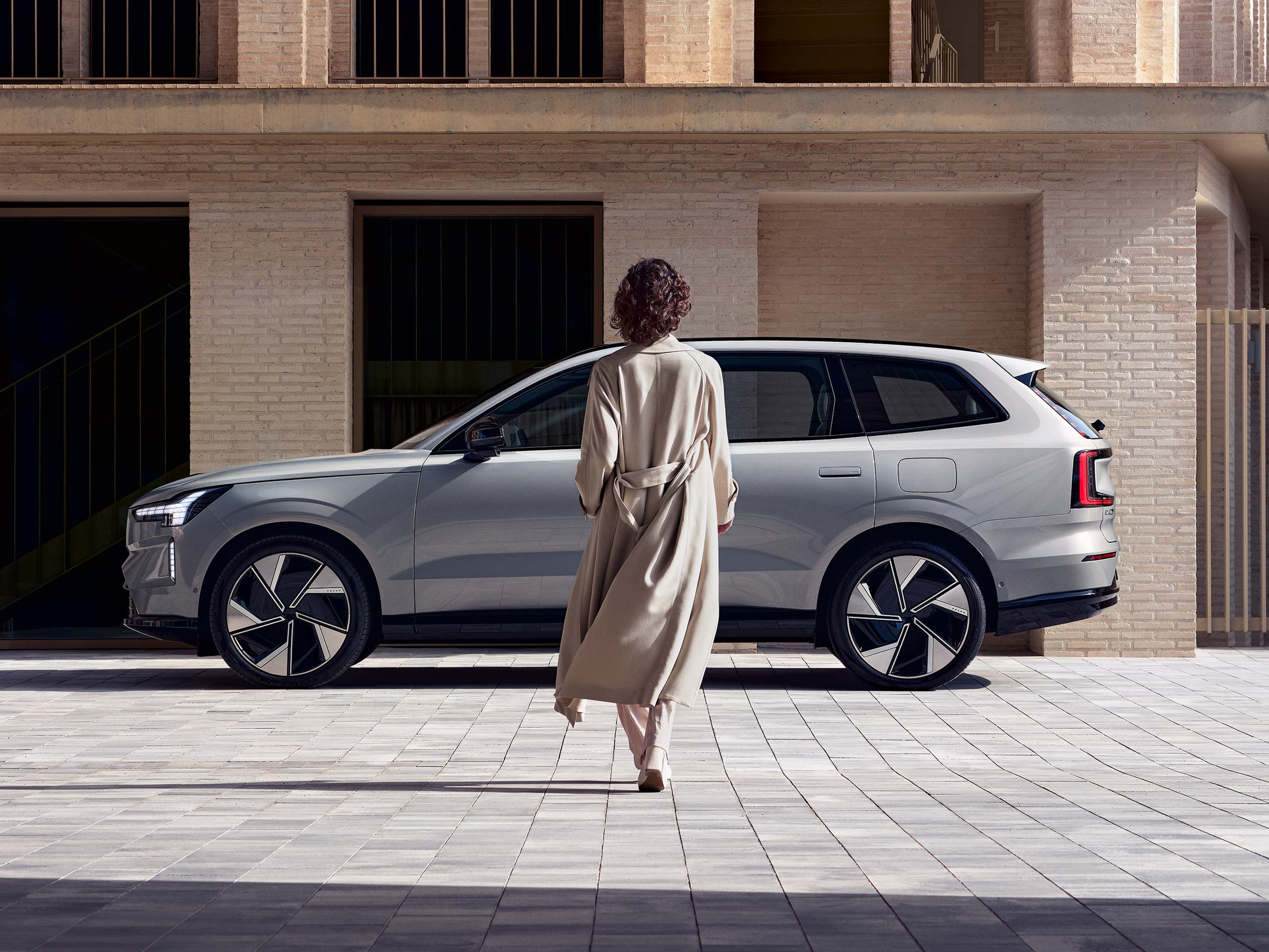 Trenssitakkiin pukeutunut nainen kävelee kohti Vapour grey -väristä Volvo EX90 täyssähköautoa. Taustalla kerrostalo.