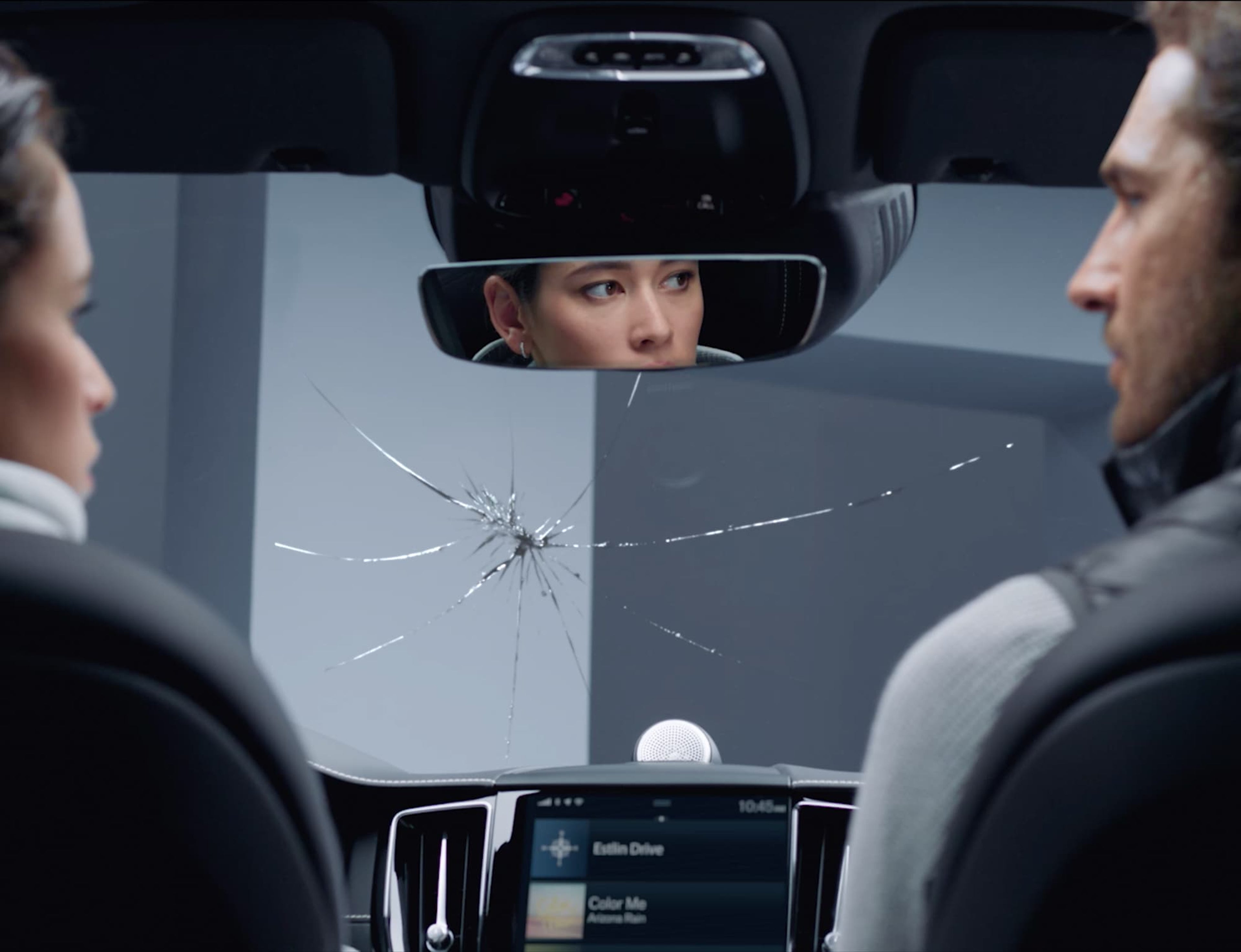 Intérieur d'une voiture avec un pare-brise fissuré, vu depuis la banquette arrière. Le conducteur est visible dans le rétroviseur.