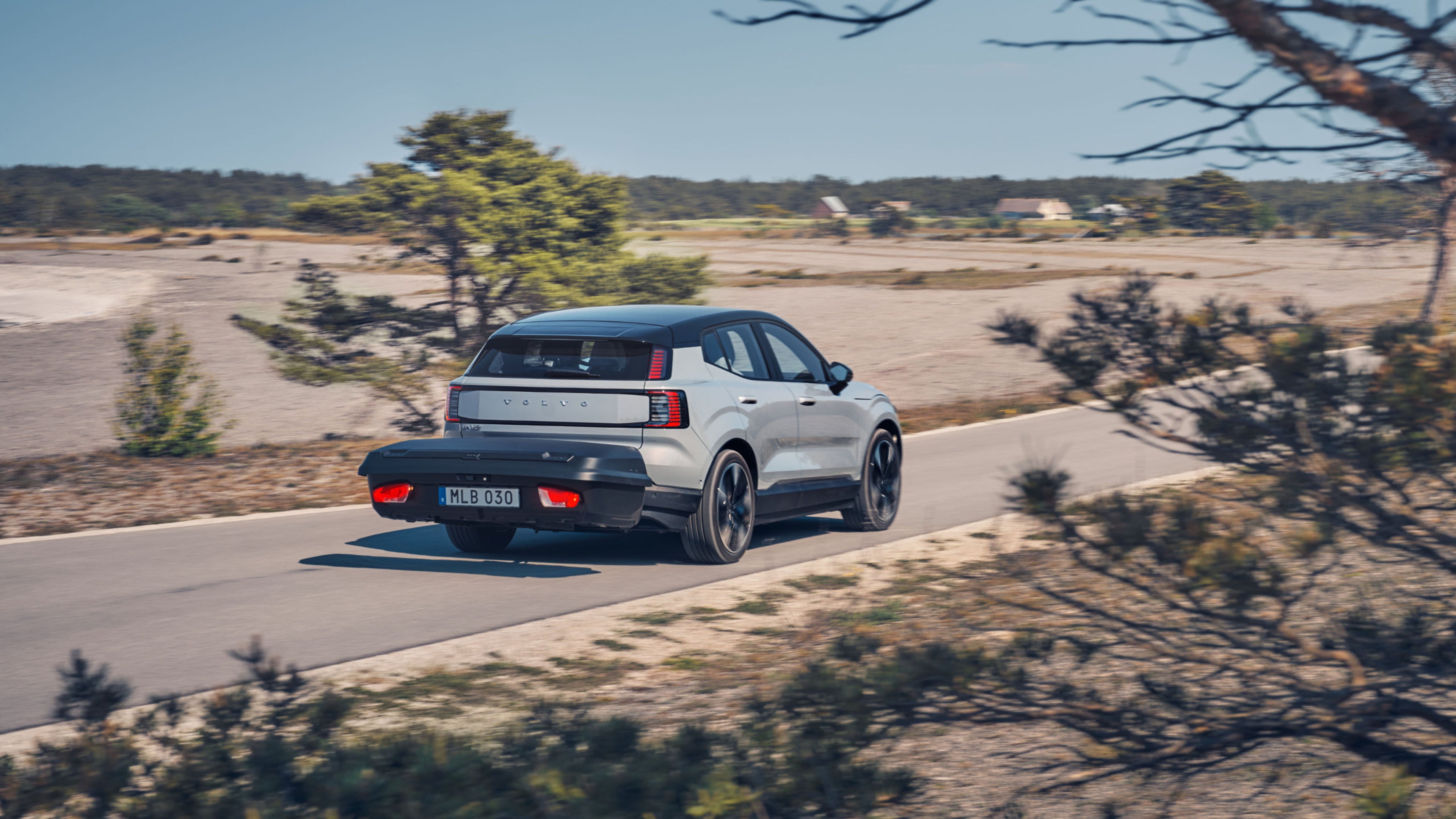 Ein grauer EX30 mit Blick auf das Heck und die Gepäck Box fährt auf einer sandigen Landstraße.