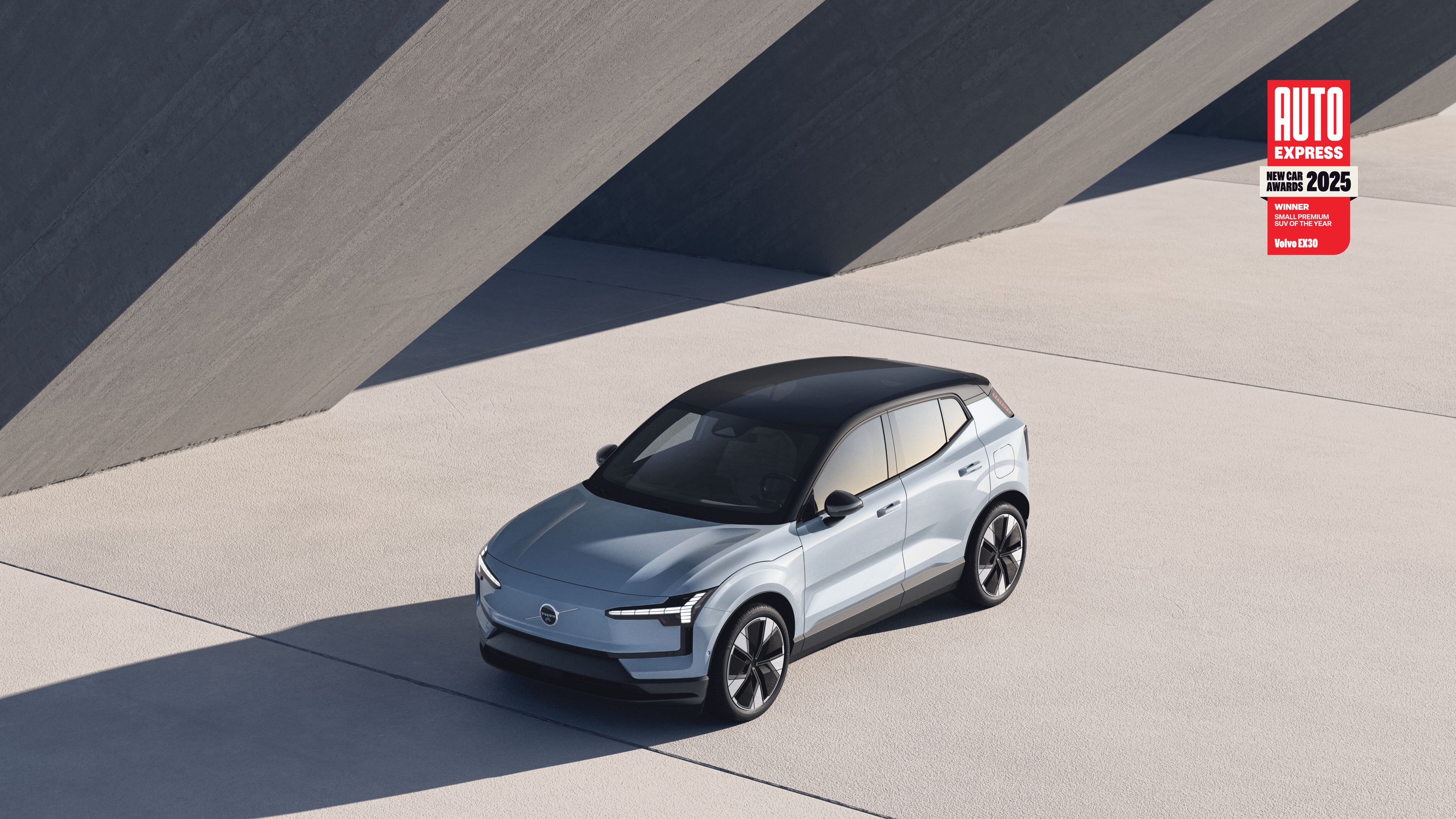 Volvo EX30 standing partly in its own shade on a road in front of a warehouse that the sun is shining on.