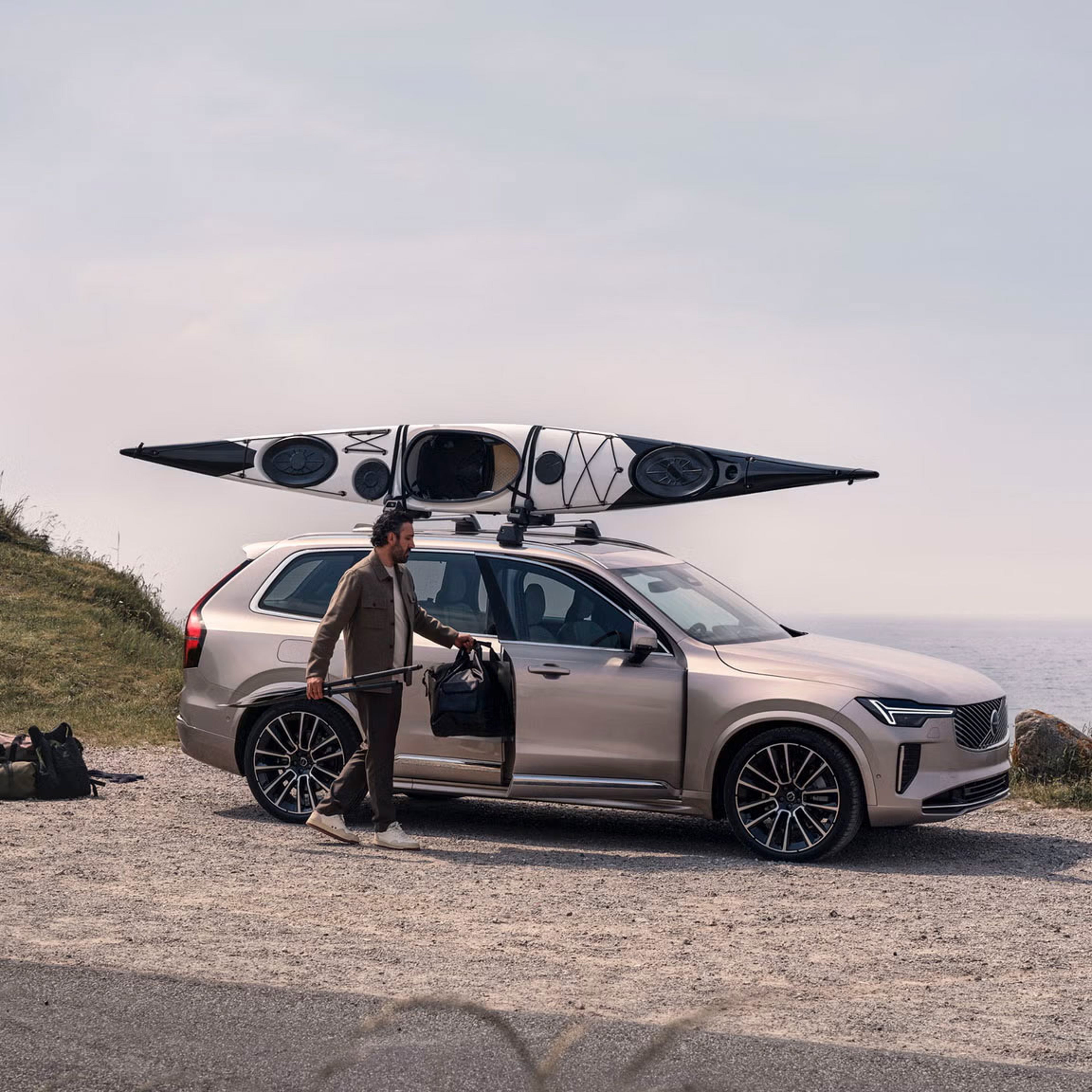 Side view of XC90 with a kayak attached to the roof, parked on gravel while someone unloads bags from open passenger door.