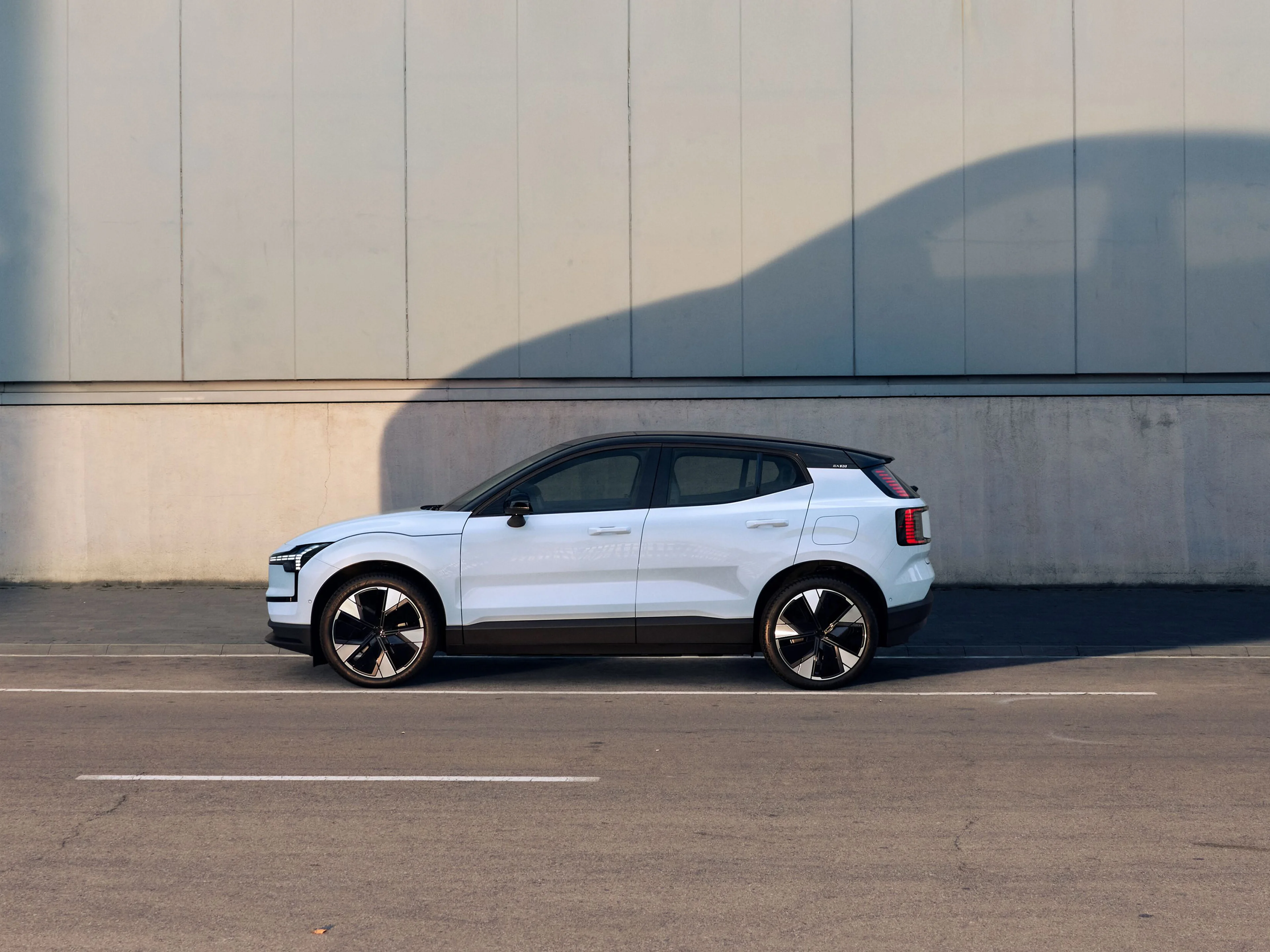 Volvo EX30 standing partly in its own shade on a road in front of a warehouse that the sun is shining on.