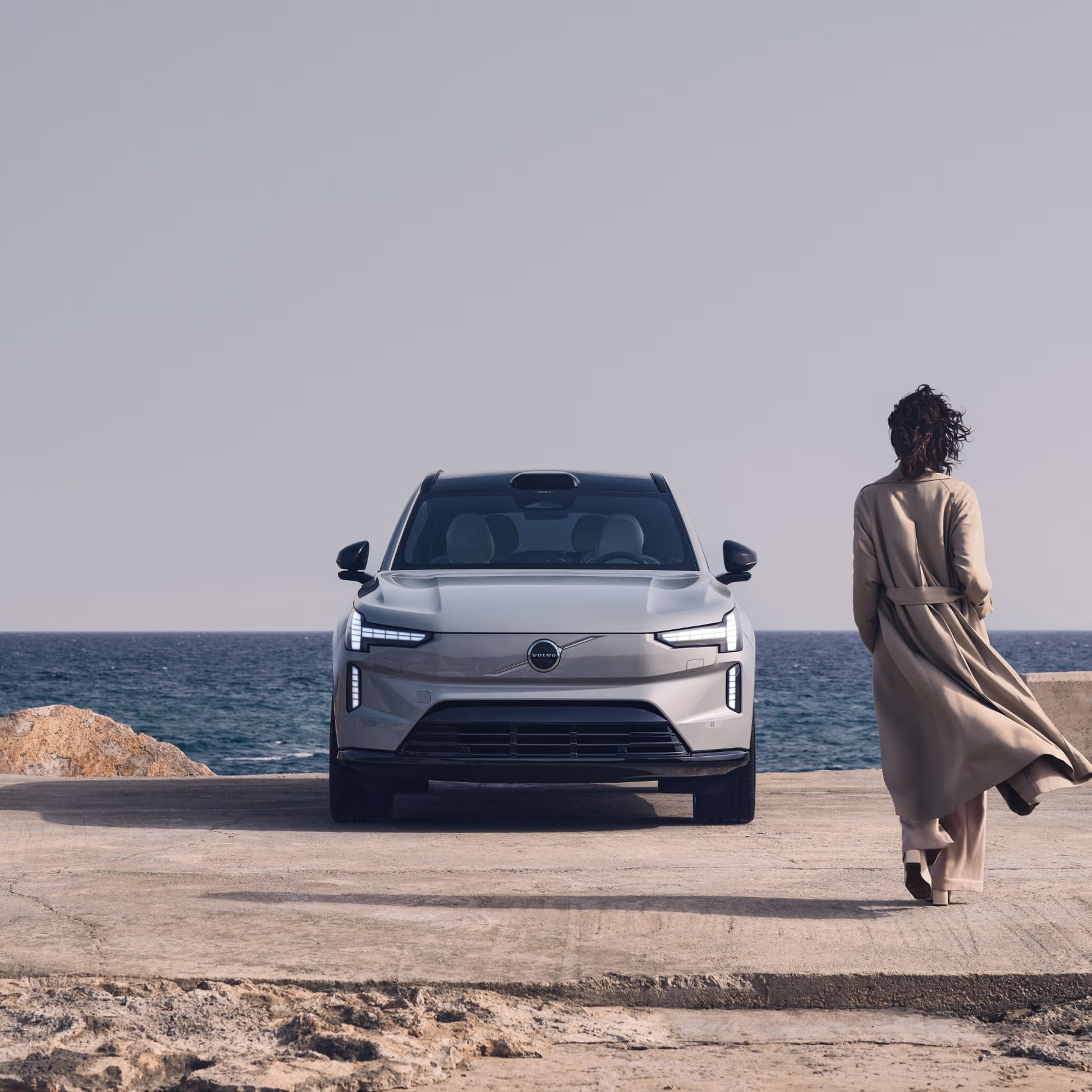 Person in trench coat walking towards EX90 parked in front of a lake on a sunny day