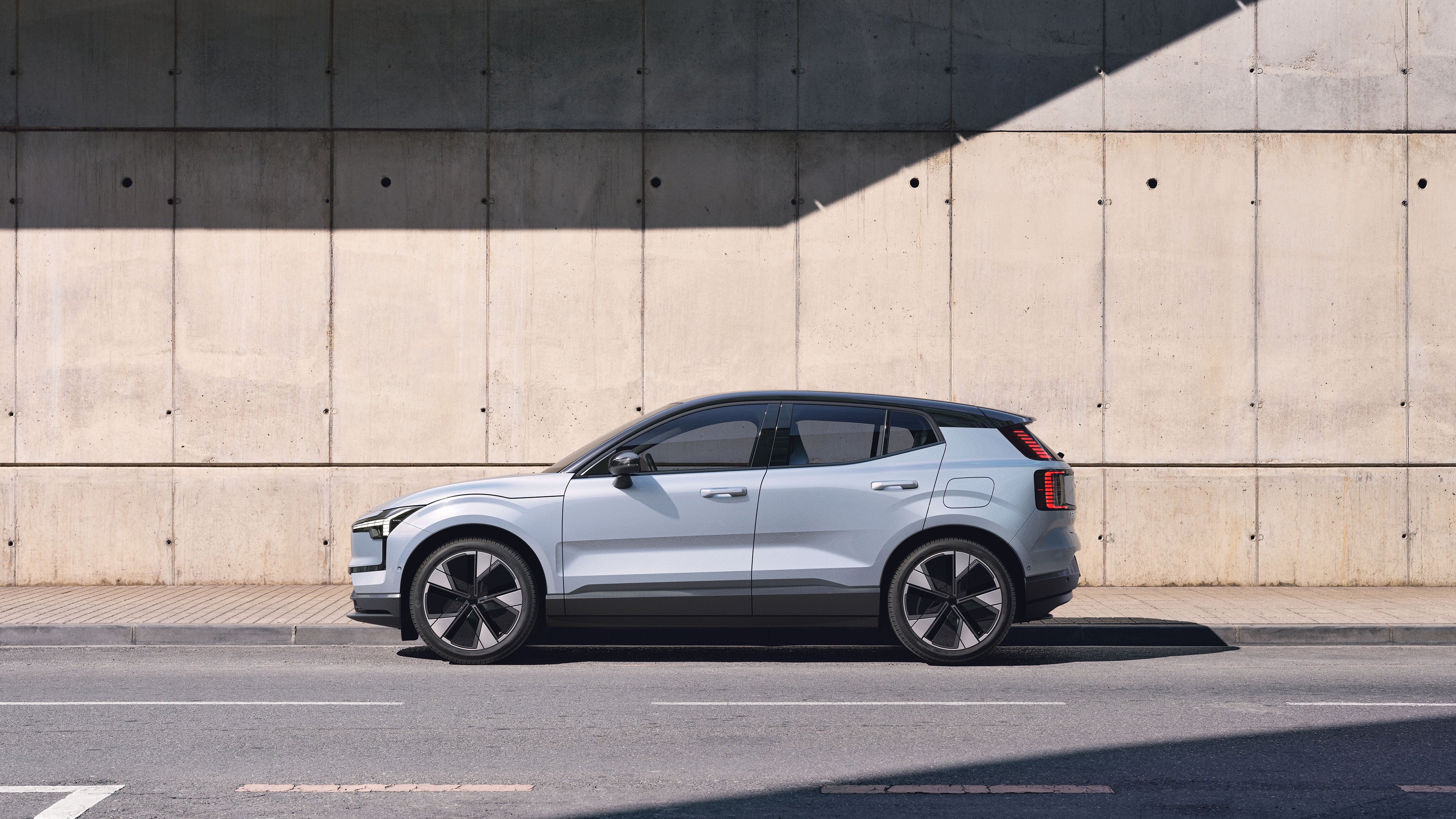 Volvo EX30 standing partly in its own shade on a road in front of a warehouse that the sun is shining on.