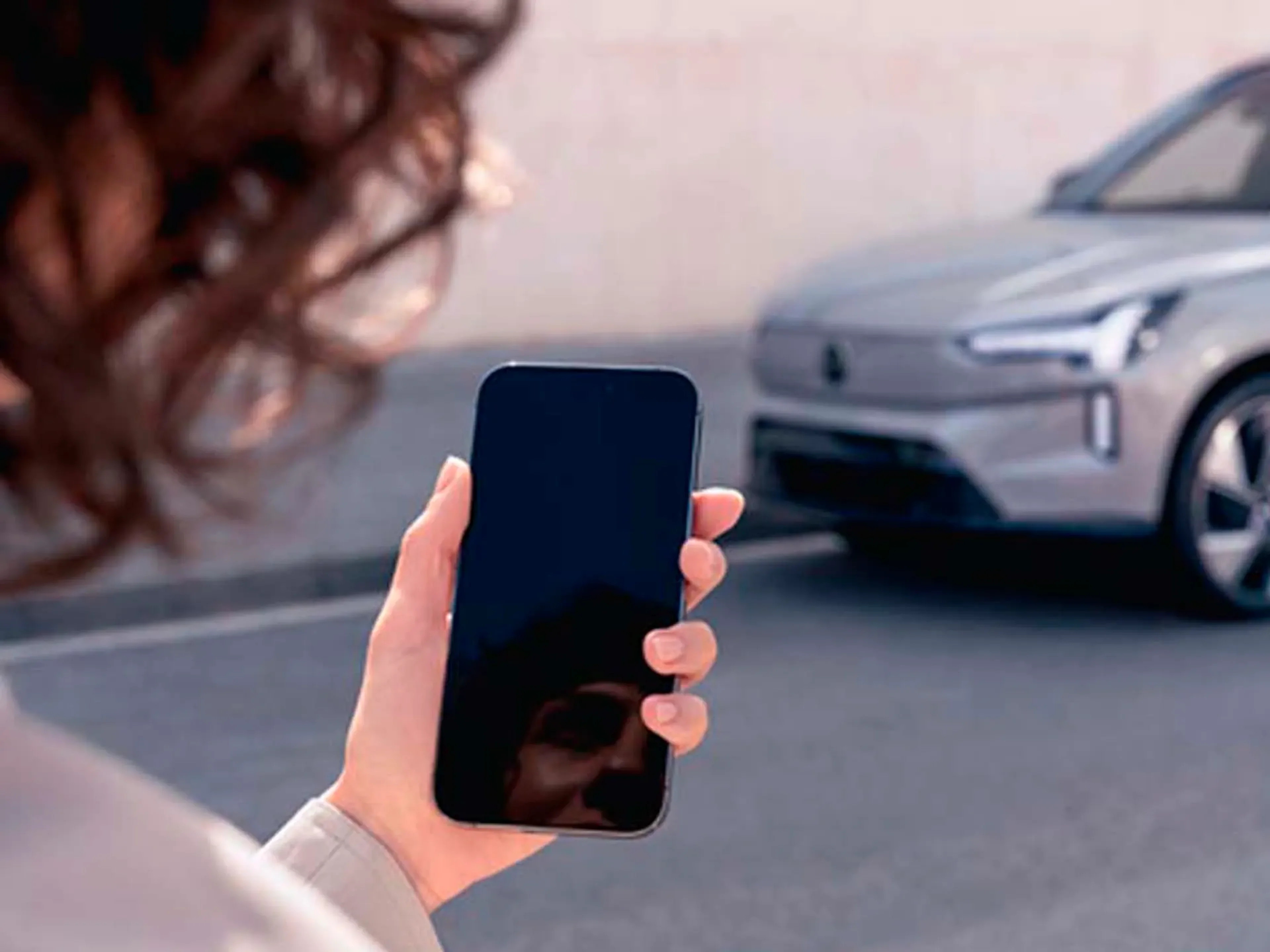 Una mujer sujetando un móvil y un vehículo Volvo al fondo.