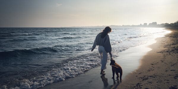 A woman walking along the beach at sunset, playing with her dog.
