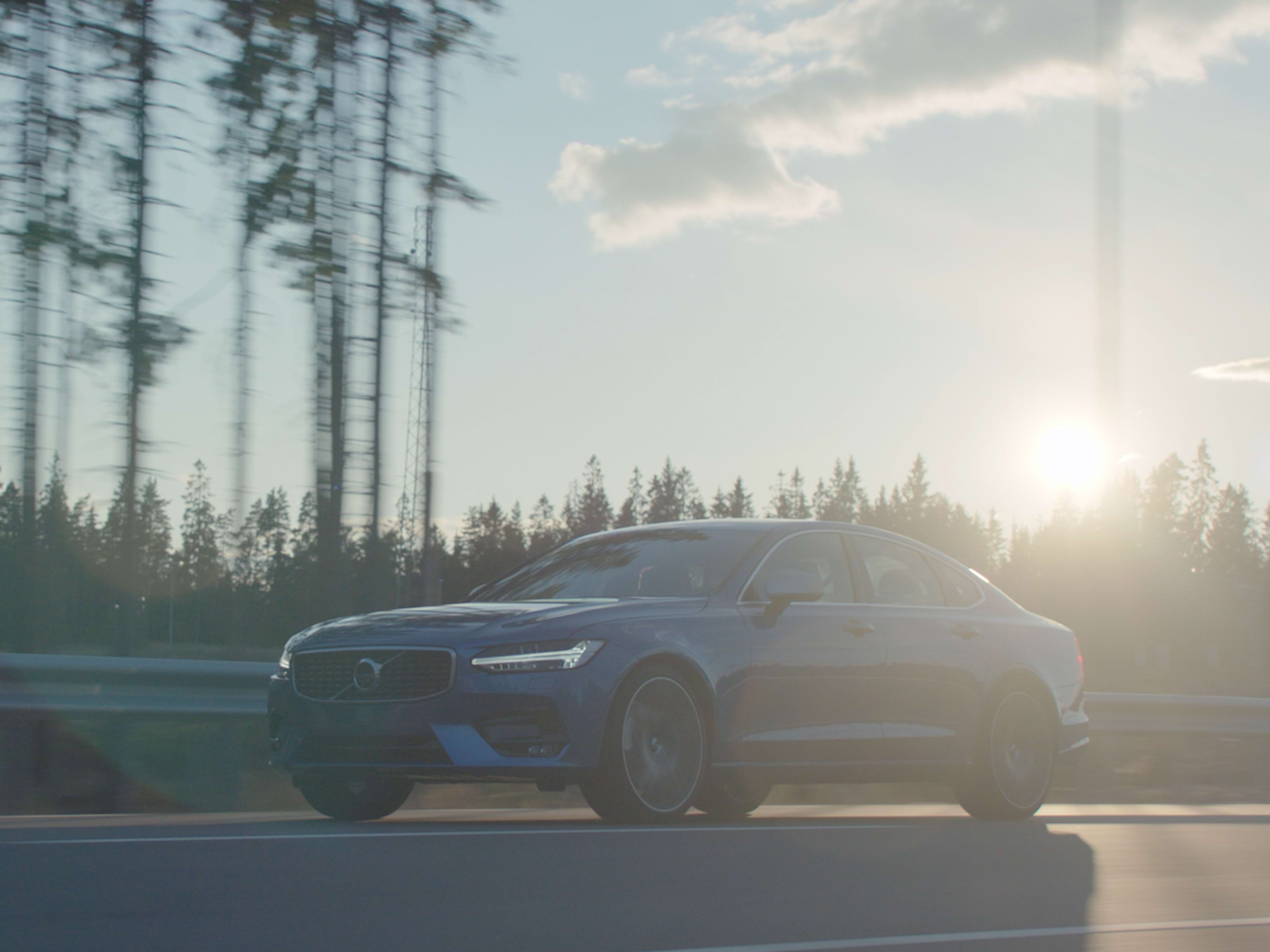 Denim kék Volvo S90 limuzin nagylátószögű felvételen, amint egy napsütéses délutánon egy erdős úton halad.