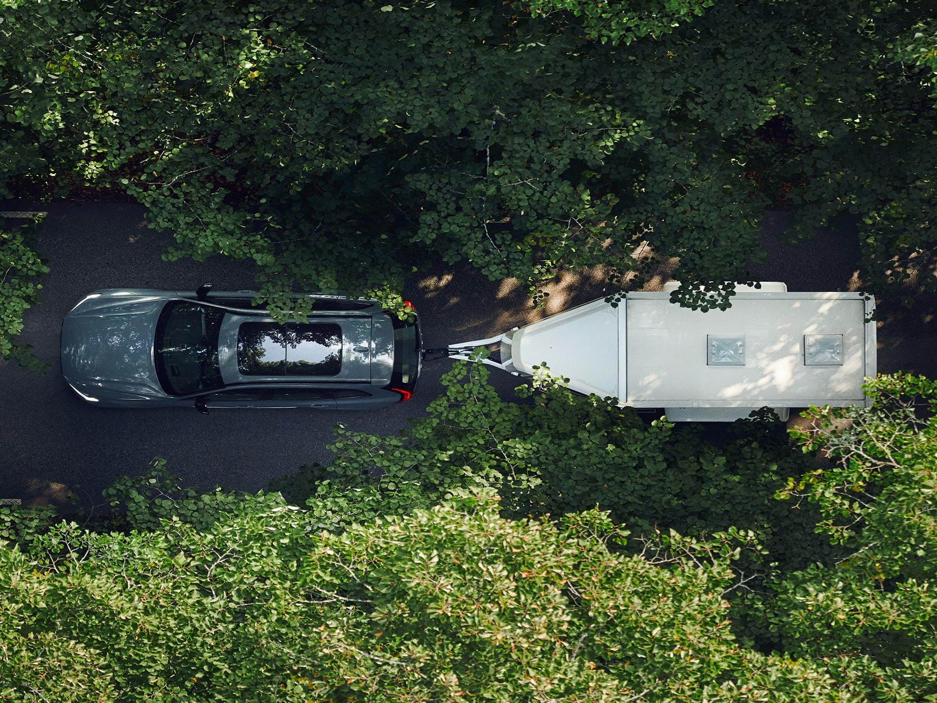 Vista de pássaro de um SUV Volvo a rebocar um atrelado branco numa estrada com folhas num dia de sol.