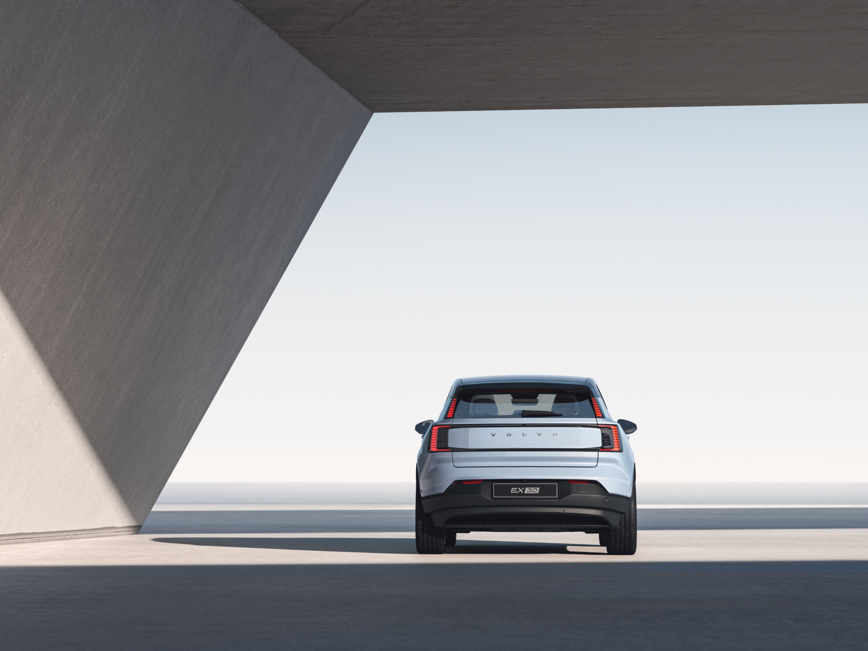 Een groothoekbeeld van de achterkant van een Volvo EX30, geparkeerd onder een betonnen overkapping in een straal zonlicht.