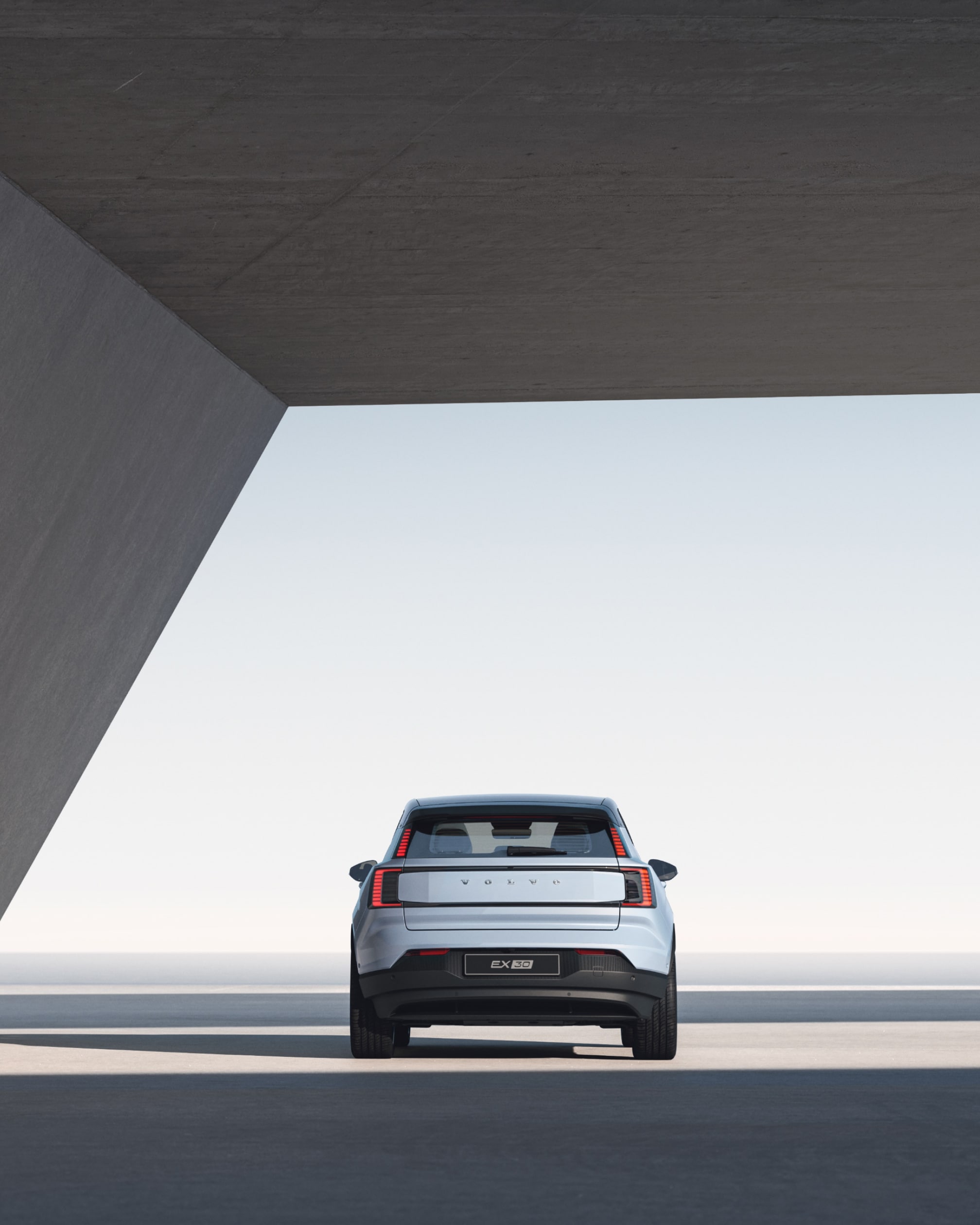 Een groothoekbeeld van de achterkant van een Volvo EX30, geparkeerd onder een betonnen overkapping in een straal zonlicht.
