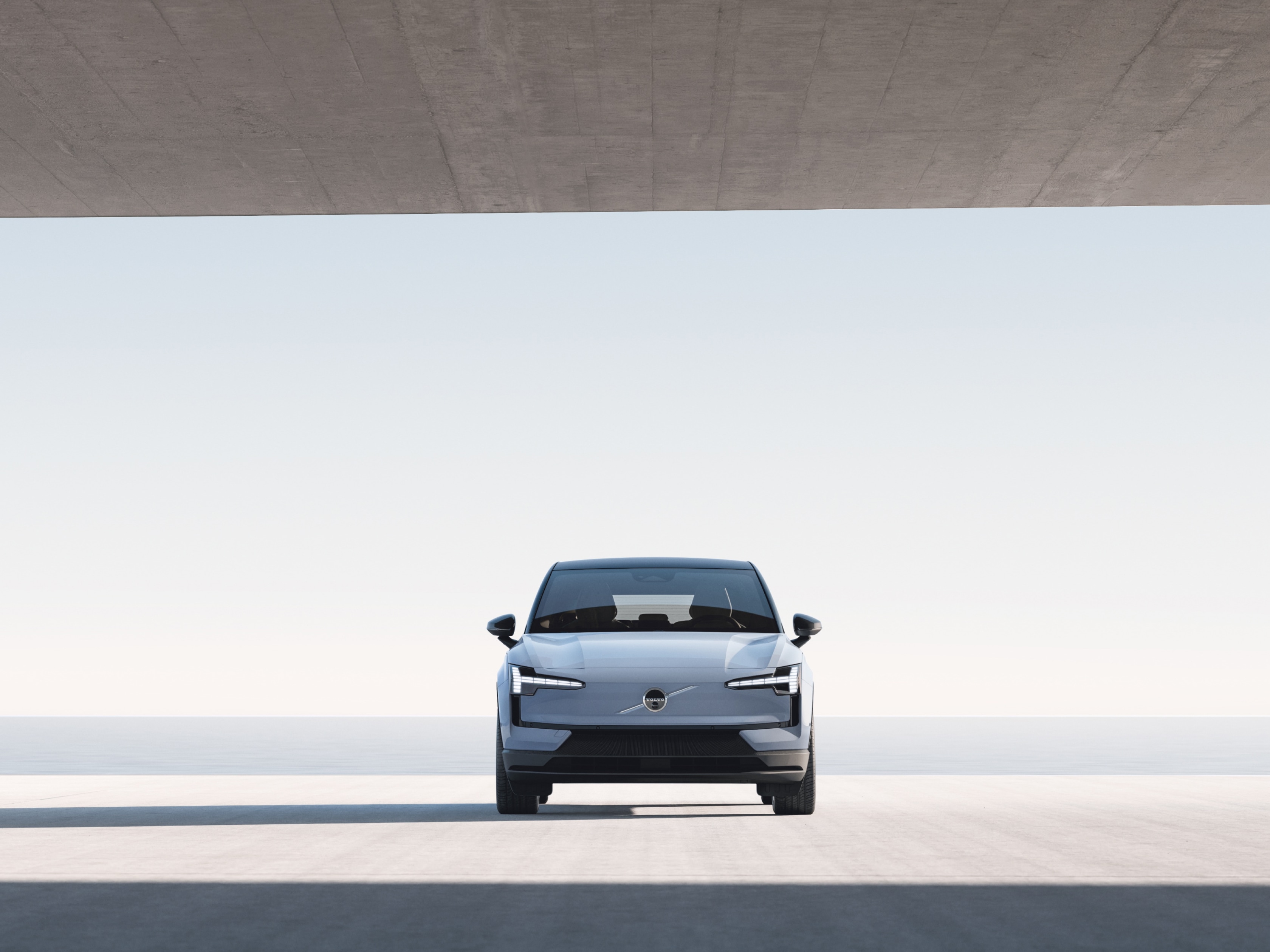 Uma imagem frontal em grande angular de um Volvo EX30 estacionado numa grande estrutura de betão com vista para a água.