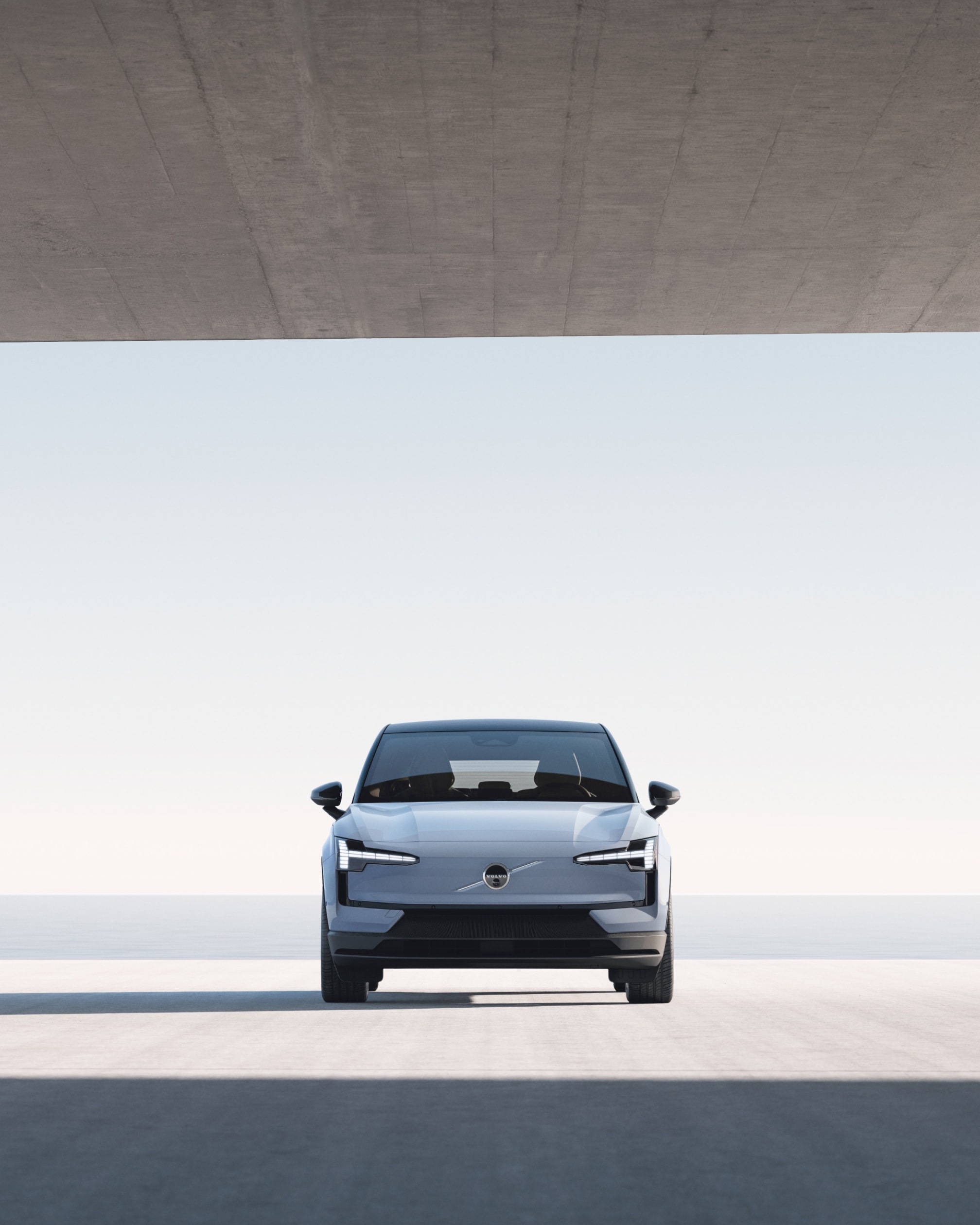 Ширококутне фронтальне зображення автомобіля Volvo EX30, припаркованого у великій бетонній споруді, з видом на воду.