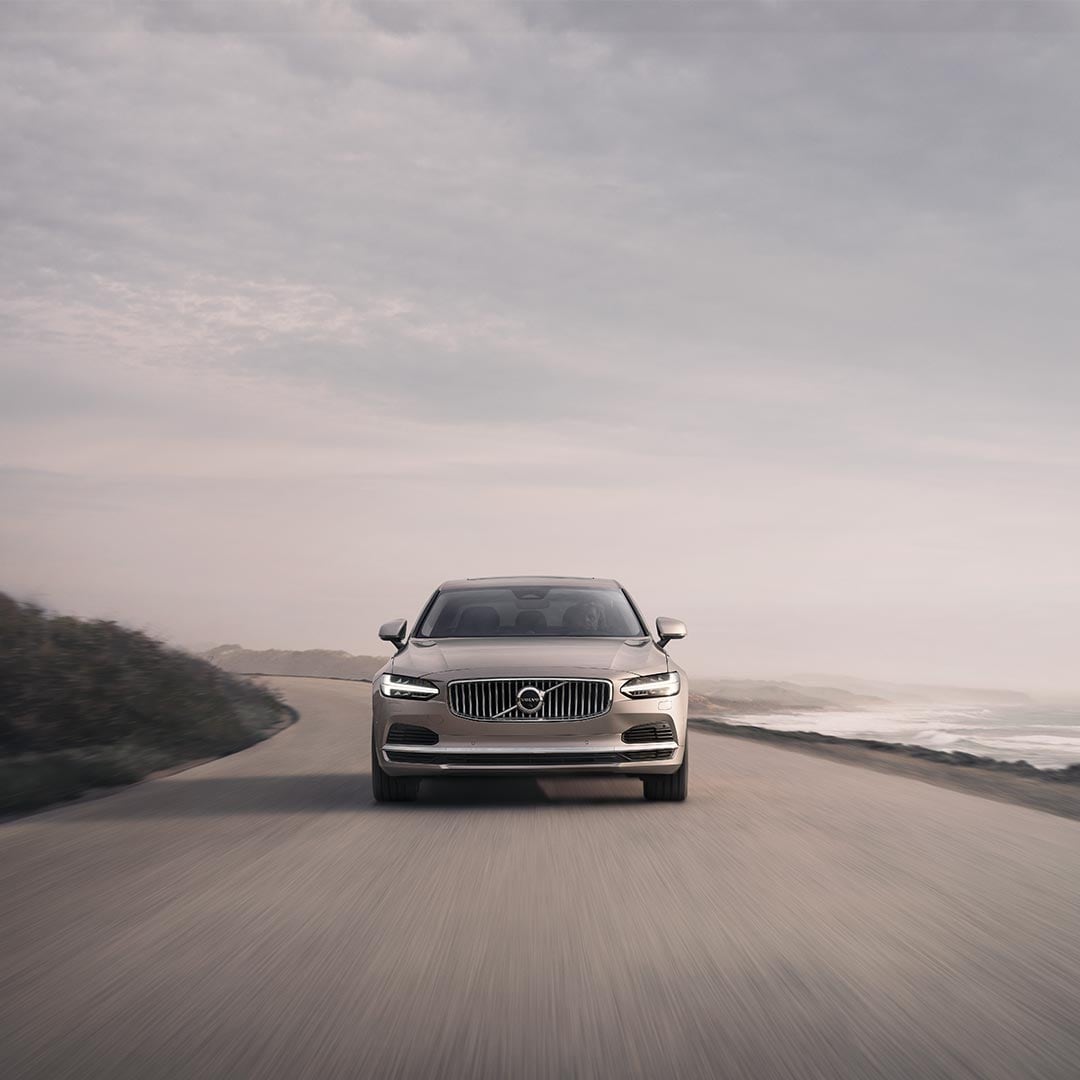 Image of the front of a Volvo S90 driving down a road next to the sea.