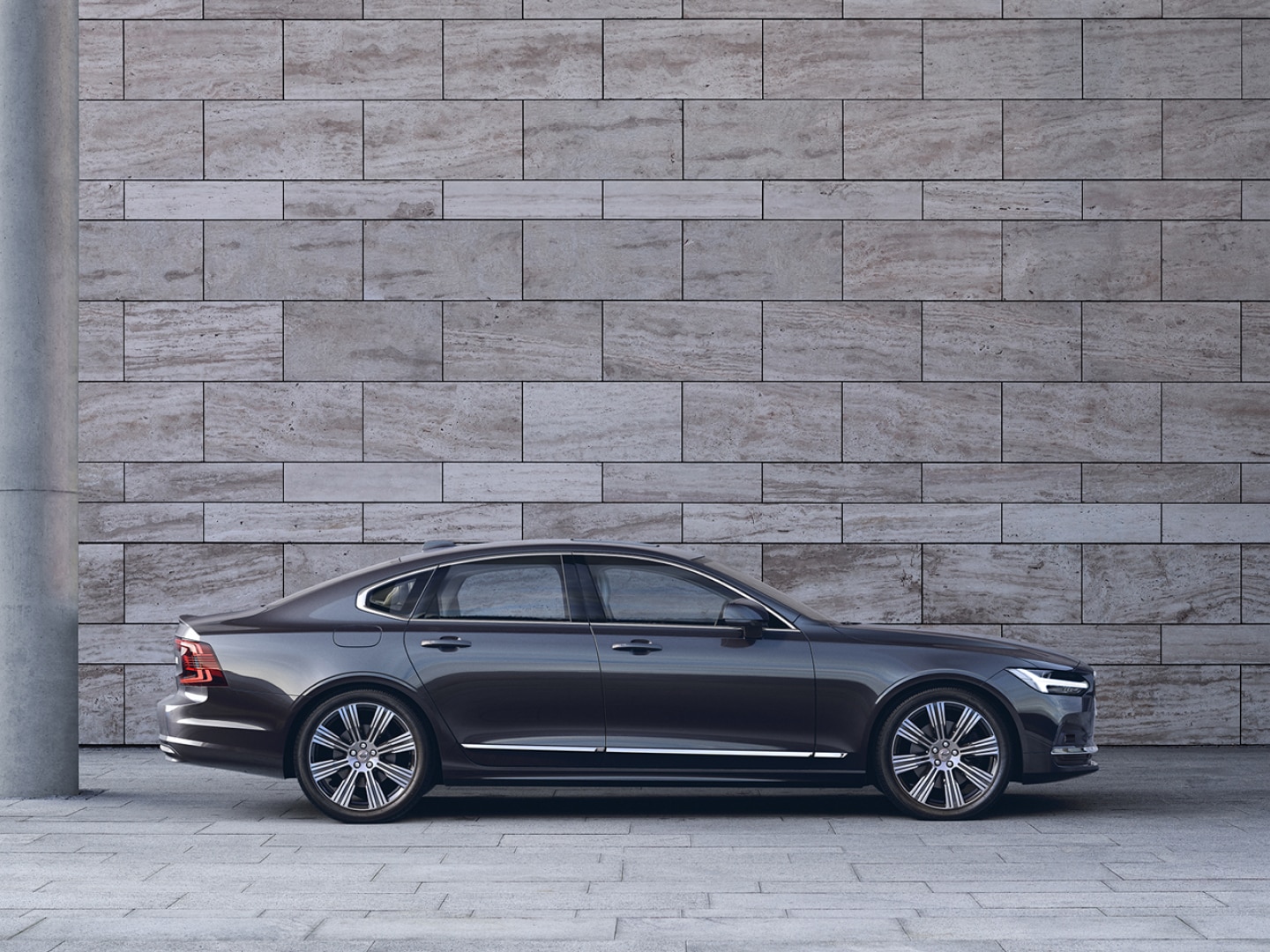 Zijaanzicht van een Volvo S90 die voor een grijze betonnen muur geparkeerd is.