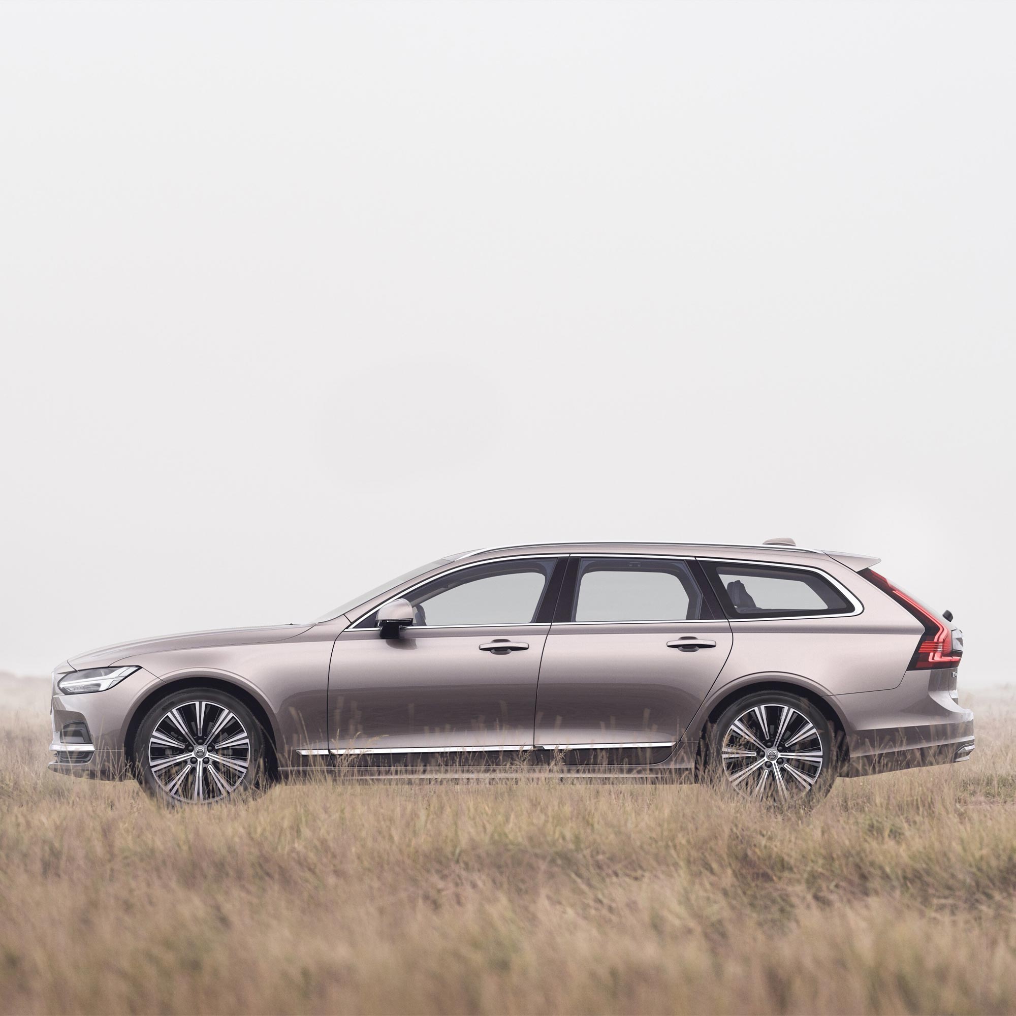 Wide shot of a Volvo V90 estate car in Bright Dusk standing parked in a misty field of soft golden grass.