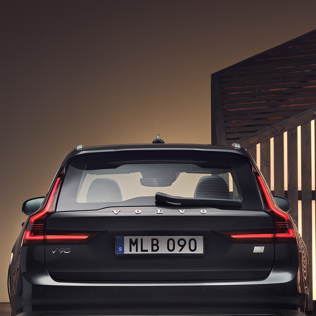 The rear exterior of a Volvo V90 Recharge, placed in a brown surrounding.