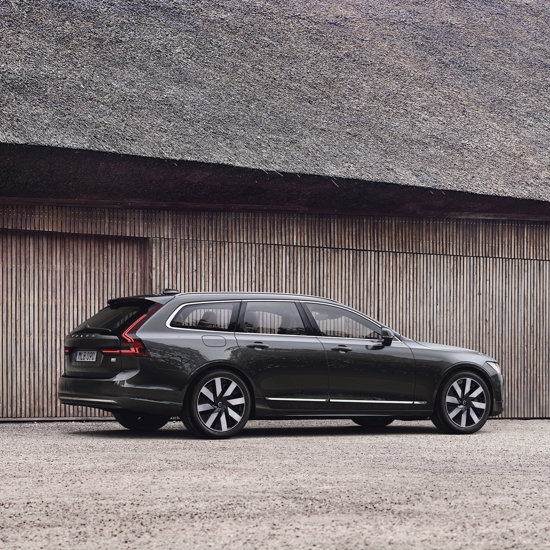 Side view of Volvo V90 Recharge parked in front of wooden structure. 