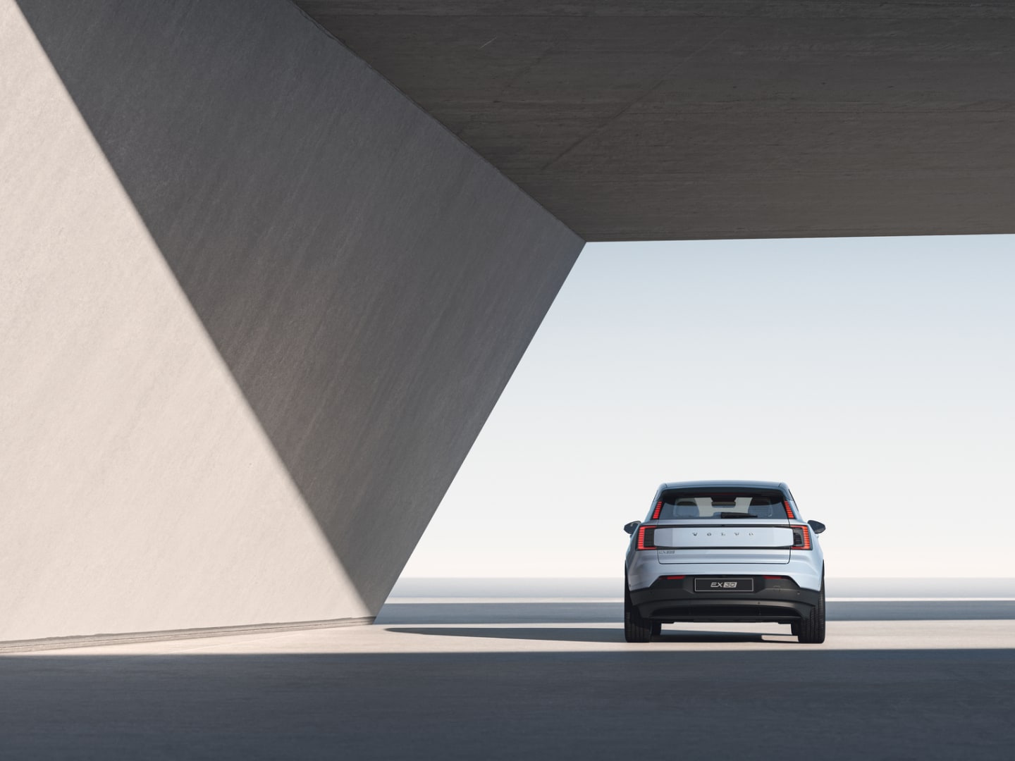 Широкоъгълно изображение на задната част на Volvo EX30, паркирано под отворена бетонна конструкция и лъчи от слънчева светлина.