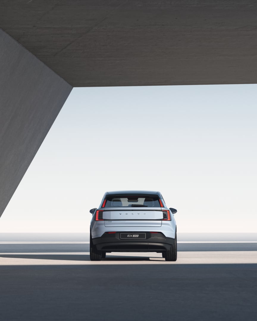 Широкоъгълно изображение на задната част на Volvo EX30, паркирано под отворена бетонна конструкция и лъчи от слънчева светлина.