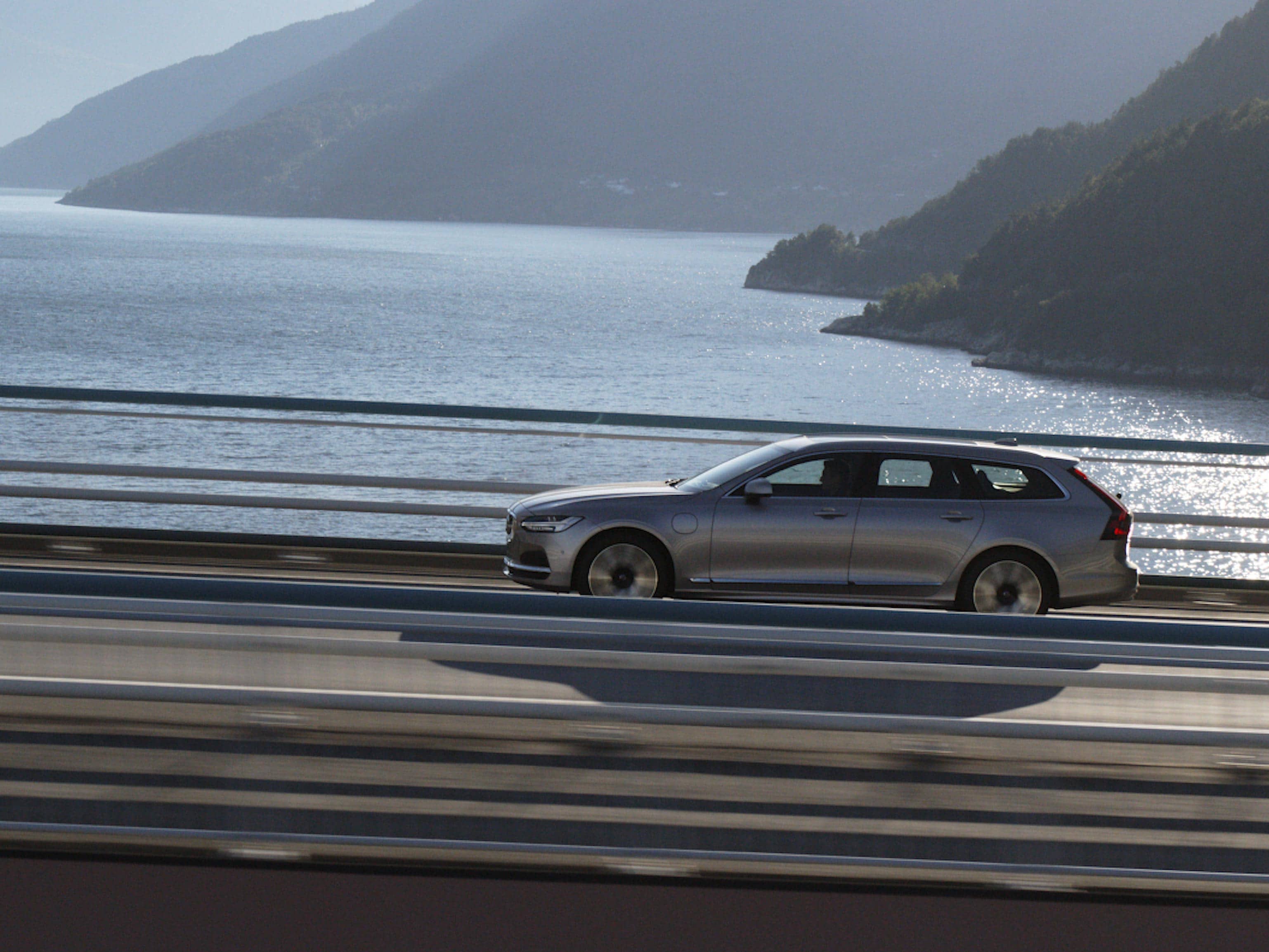 Širokoúhlý pohled na kombi Volvo V90 v zlatavě stříbrné barvě Bright Dusk jedoucí po horské silnici nad třpytivým fjordem.