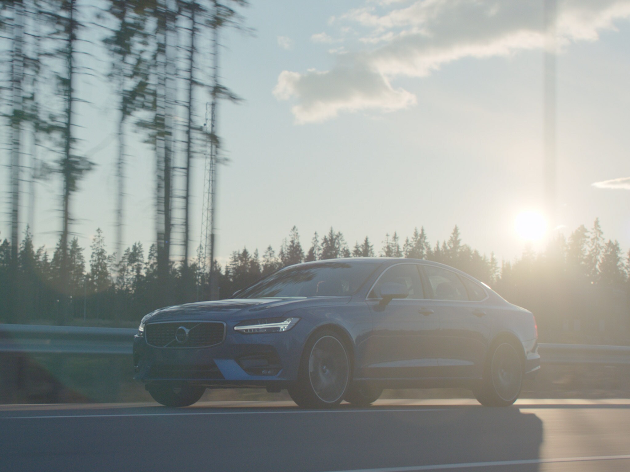 Širokoúhlý pohled na sedan Volvo S90 v tmavě modré barvě Denim Blue jedoucí po lesní silnici za slunečného dne.