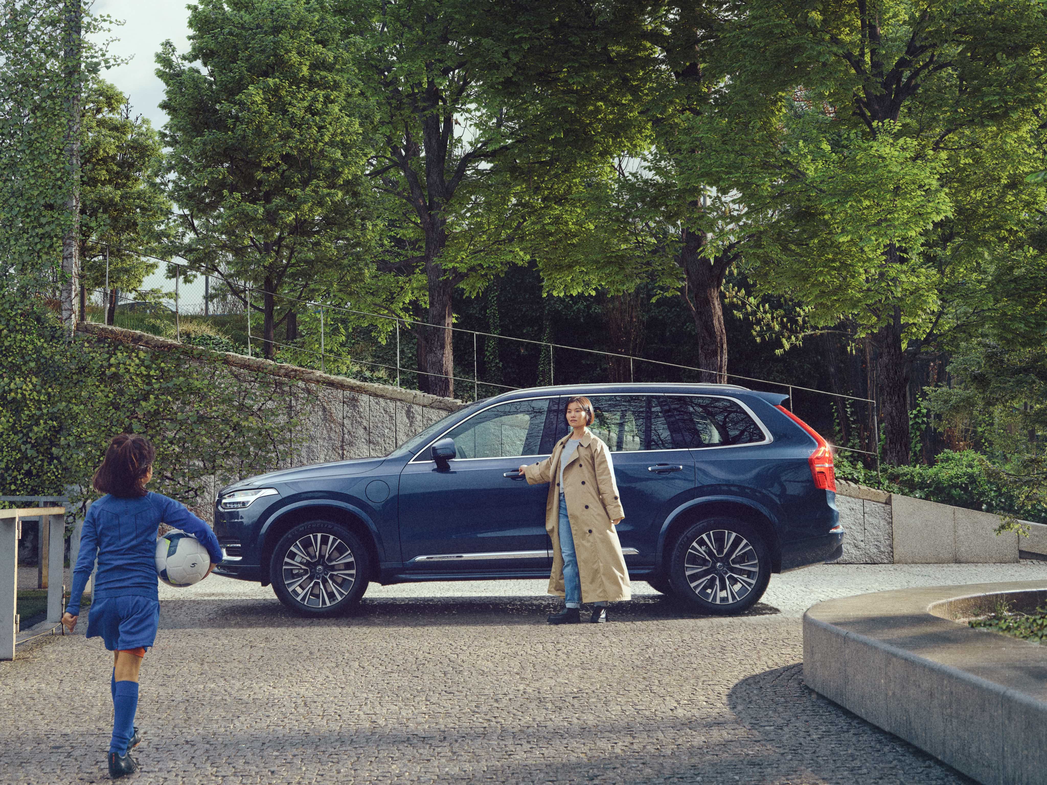 Eine Mutter steht neben ihrem Volvo XC90 in Denim Blue, während sich ihre Tochter nähert, die zum Fussballtraining angezogen ist und einen Ball in der Hand hält.