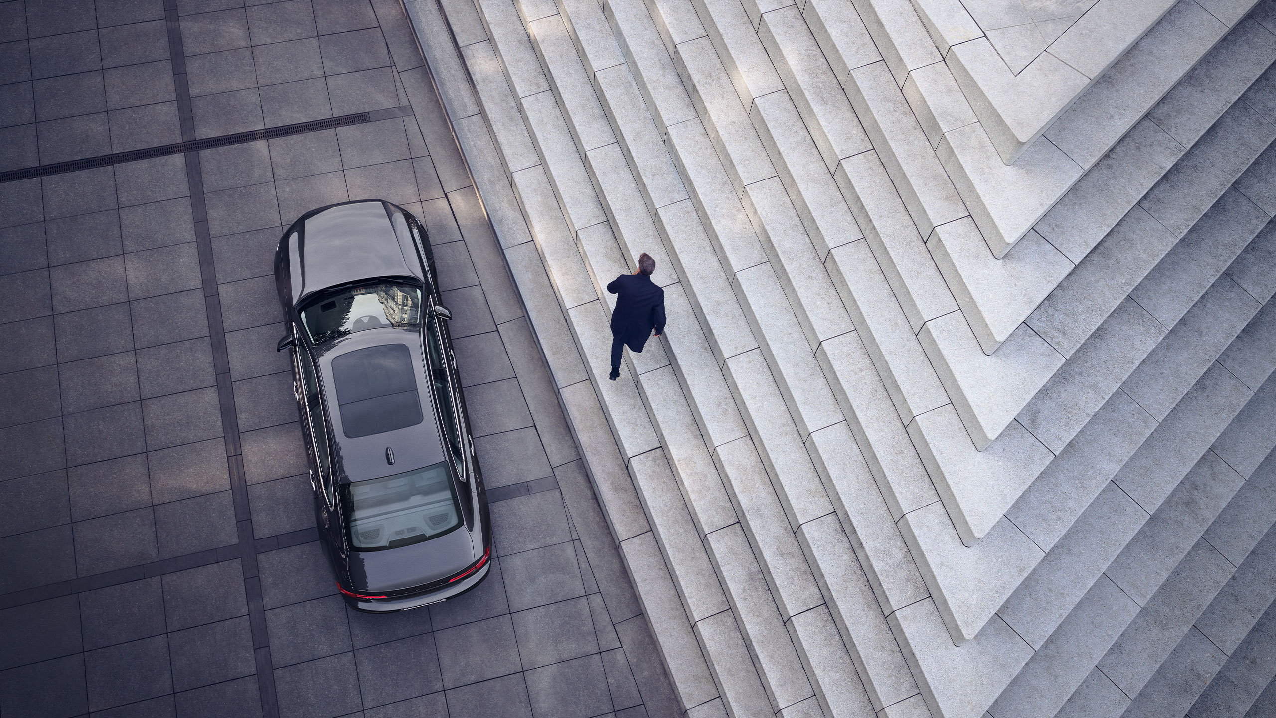 Ein Mann, der seinen Volvo S90 neben einer grafischen Marmortreppe abstellt, aus der Vogelperspektive.