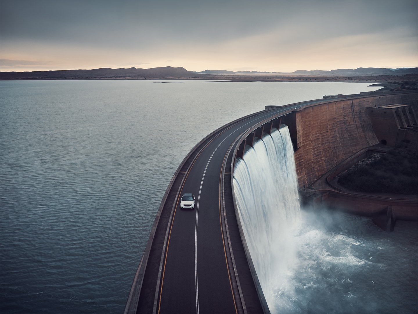 A Volvo SUV driving on a road across a dam holding a reservoir.