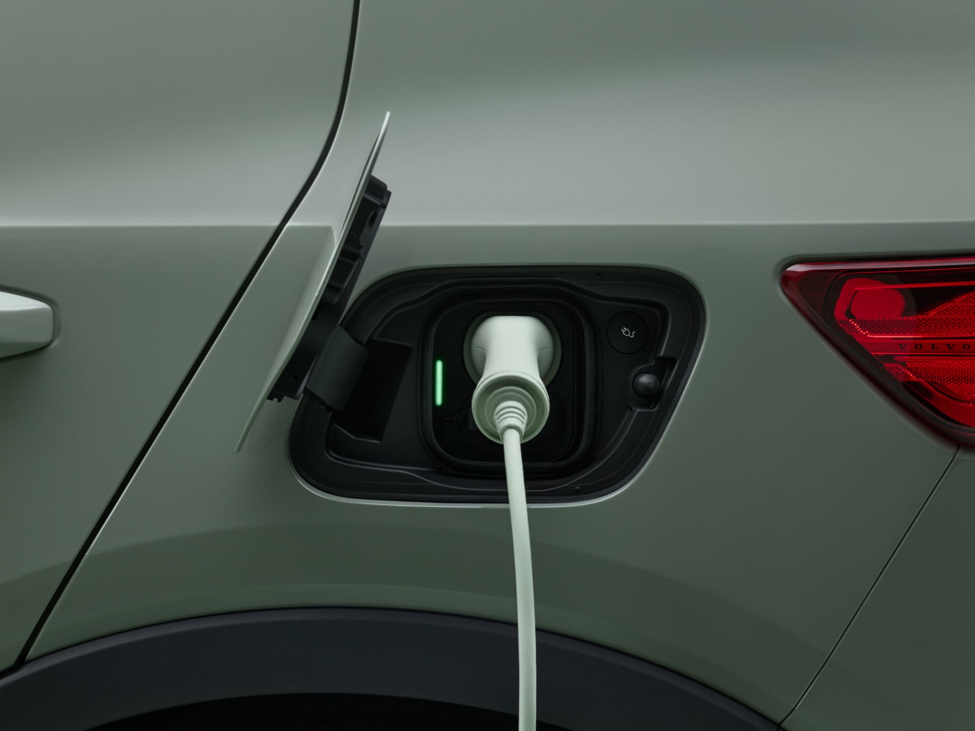 Close-up of driver's hand using a Plugsurfing charging key to activate charging station. In the middle ground, a left rear quarter-view of a new Volvo XC40 Recharge Pure Electric SUV, with charging plug inserted into car's charging port.