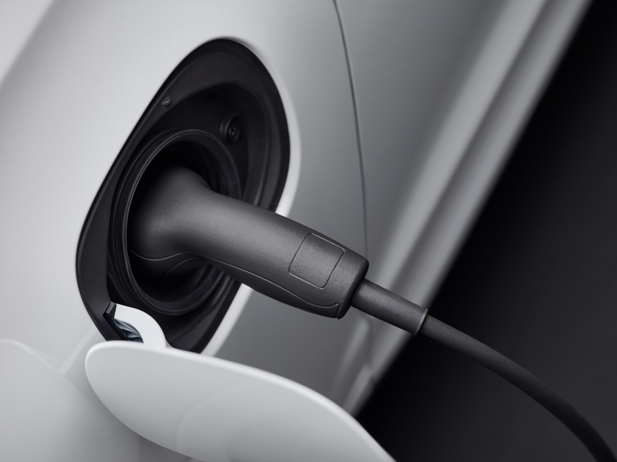 A white Volvo car's charging cap being opened and charger plugged in.