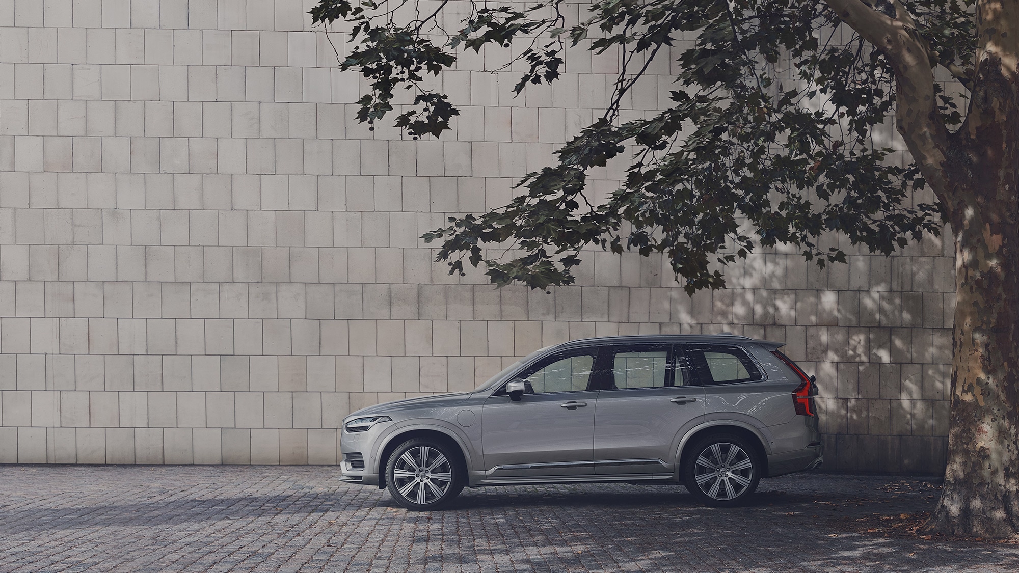 Een grijze Volvo XC90 Recharge staat geparkeerd in de schaduw onder een boom op een binnenplaats.