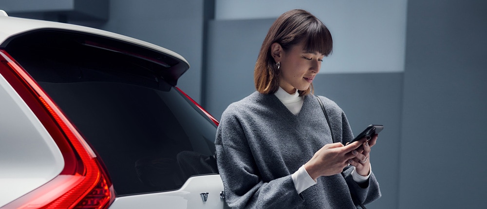 Vrouw leunt tegen de achterkant van een Volvo en bekijkt haar smartphone.