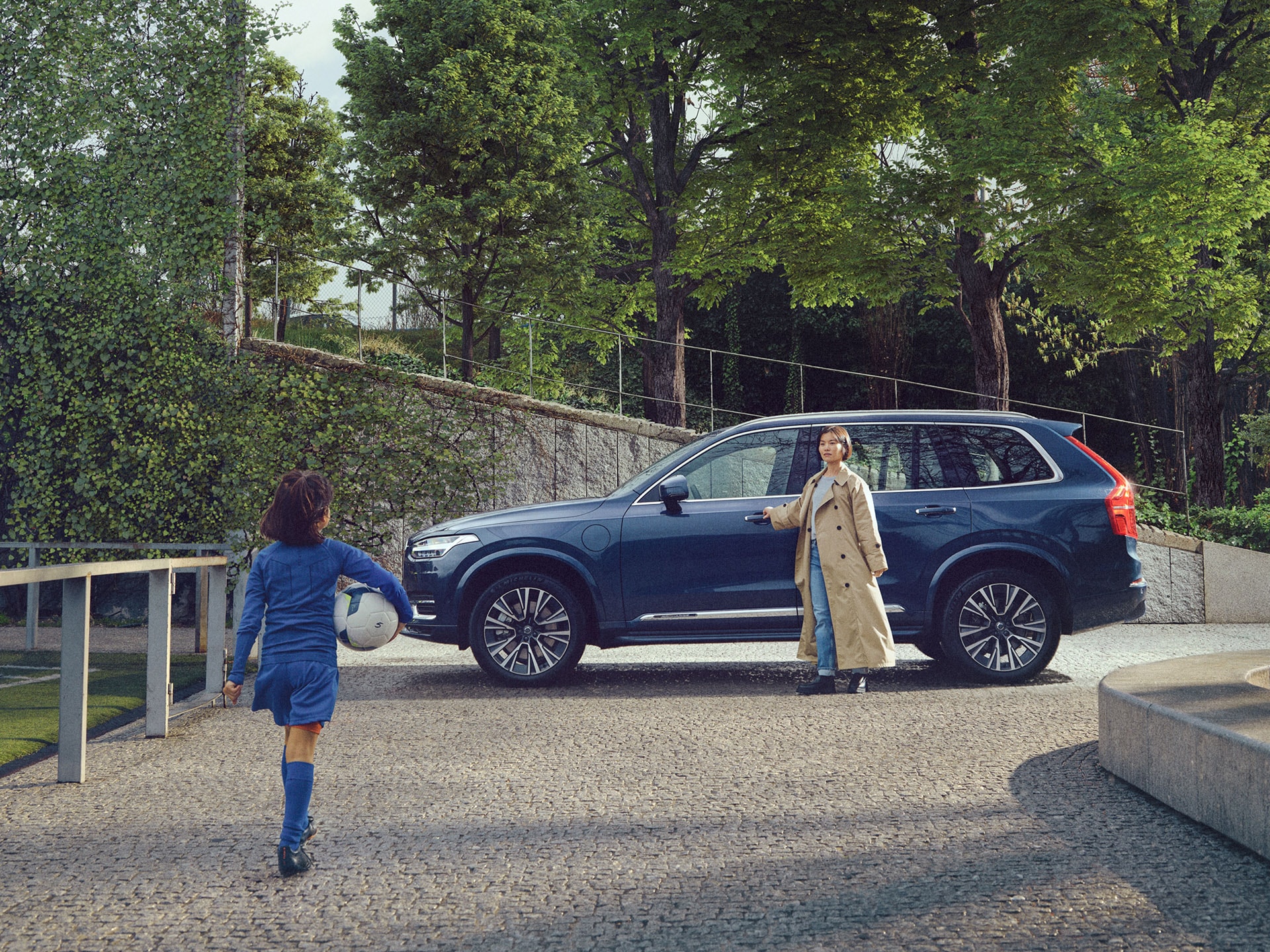 Een vrouw in een lange overjas wacht bij een blauwe Volvo XC90 op haar kind, dat een voetbal onder de arm heeft.