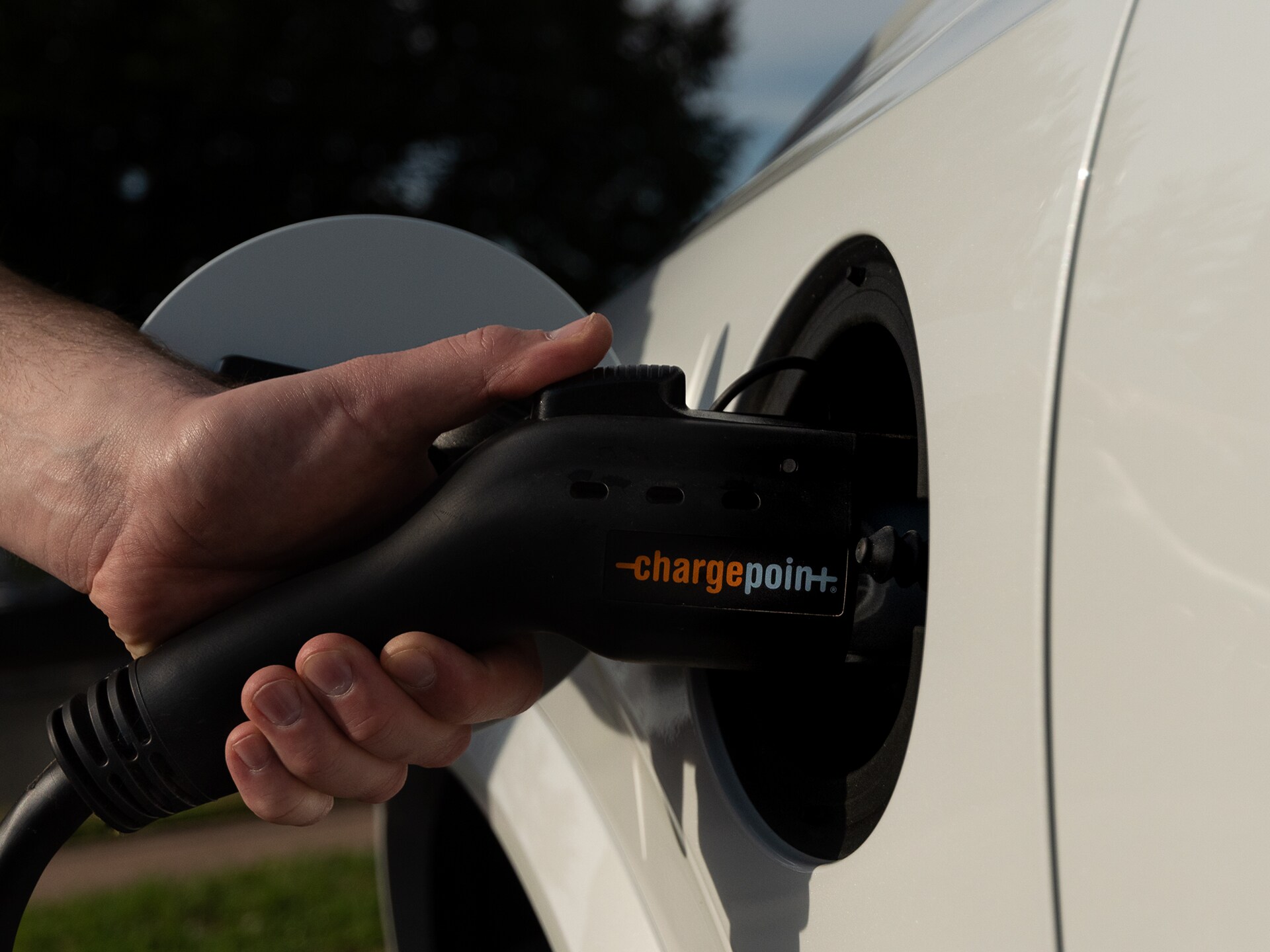 Close-up of driver's hand using a Plugsurfing charging key to activate charging station. In the middle ground, a left rear quarter-view of a new Volvo XC40 Recharge Pure Electric SUV, with charging plug inserted into car's charging port.