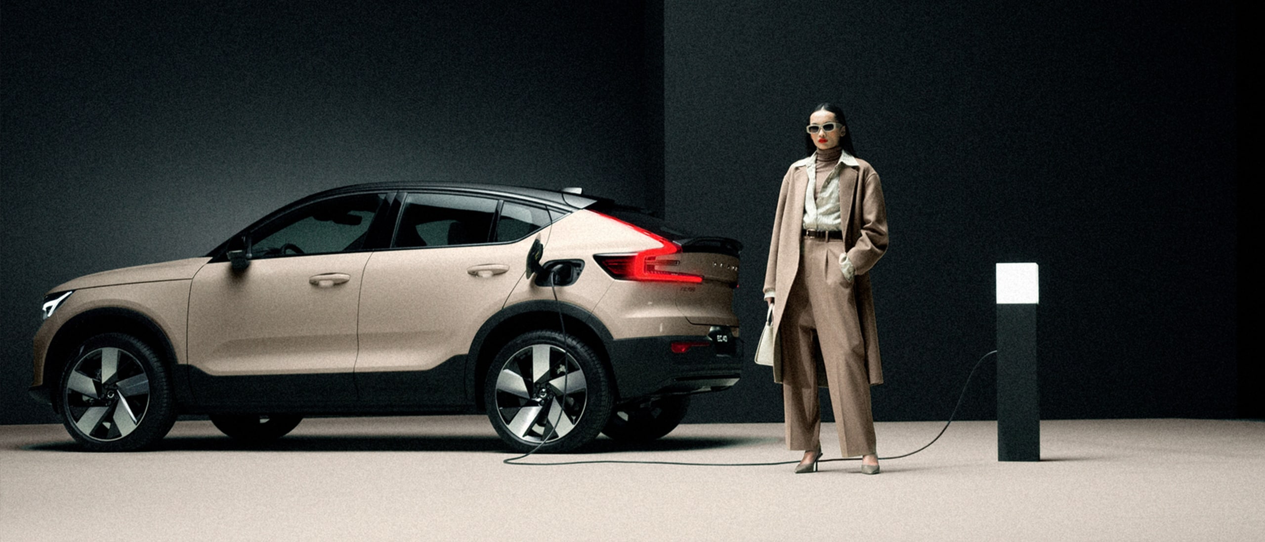Muodikkaasti pukeutunut nainen odottaa Sand Dune -värisen Volvo EC40 auton latautumista.