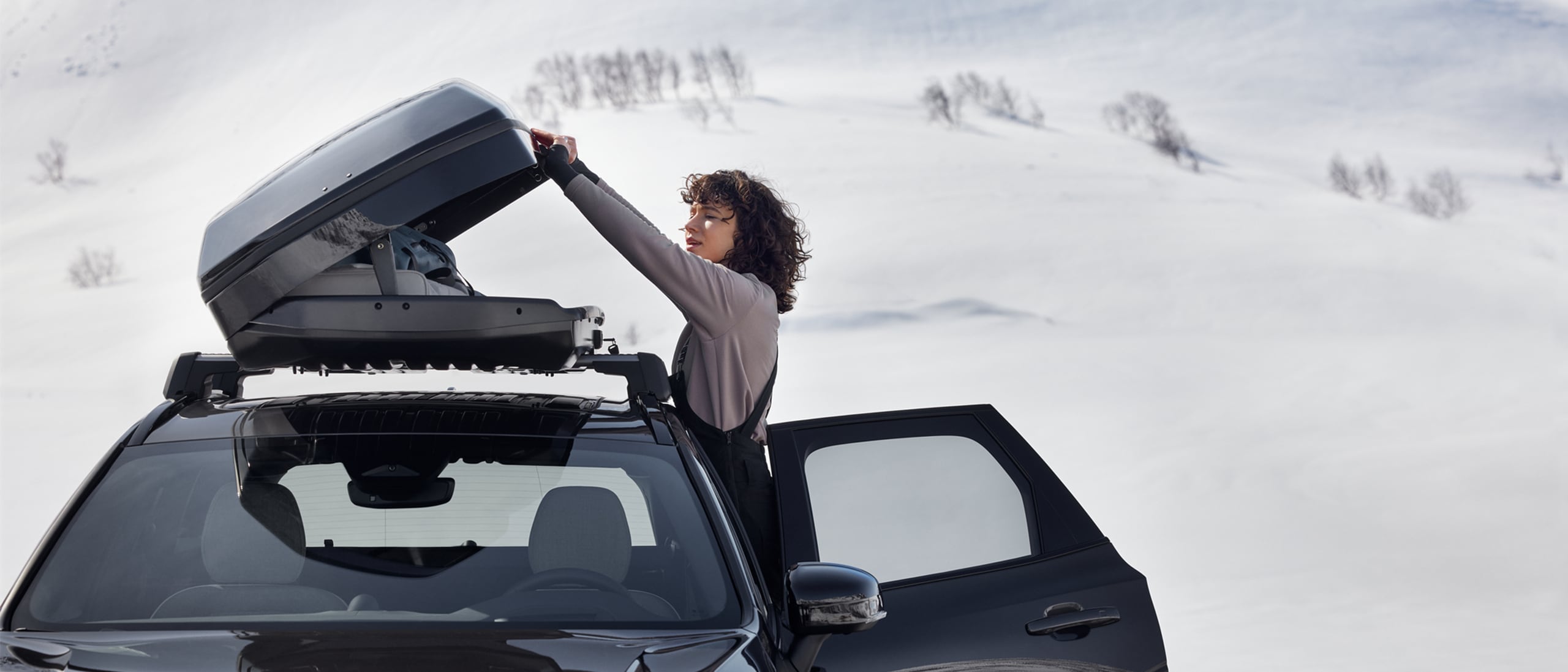Nainen avaa Platinum Grey -värisen Volvo XC60 auton katolla olevaa kattolaatikkoa lumisessa maisemassa.