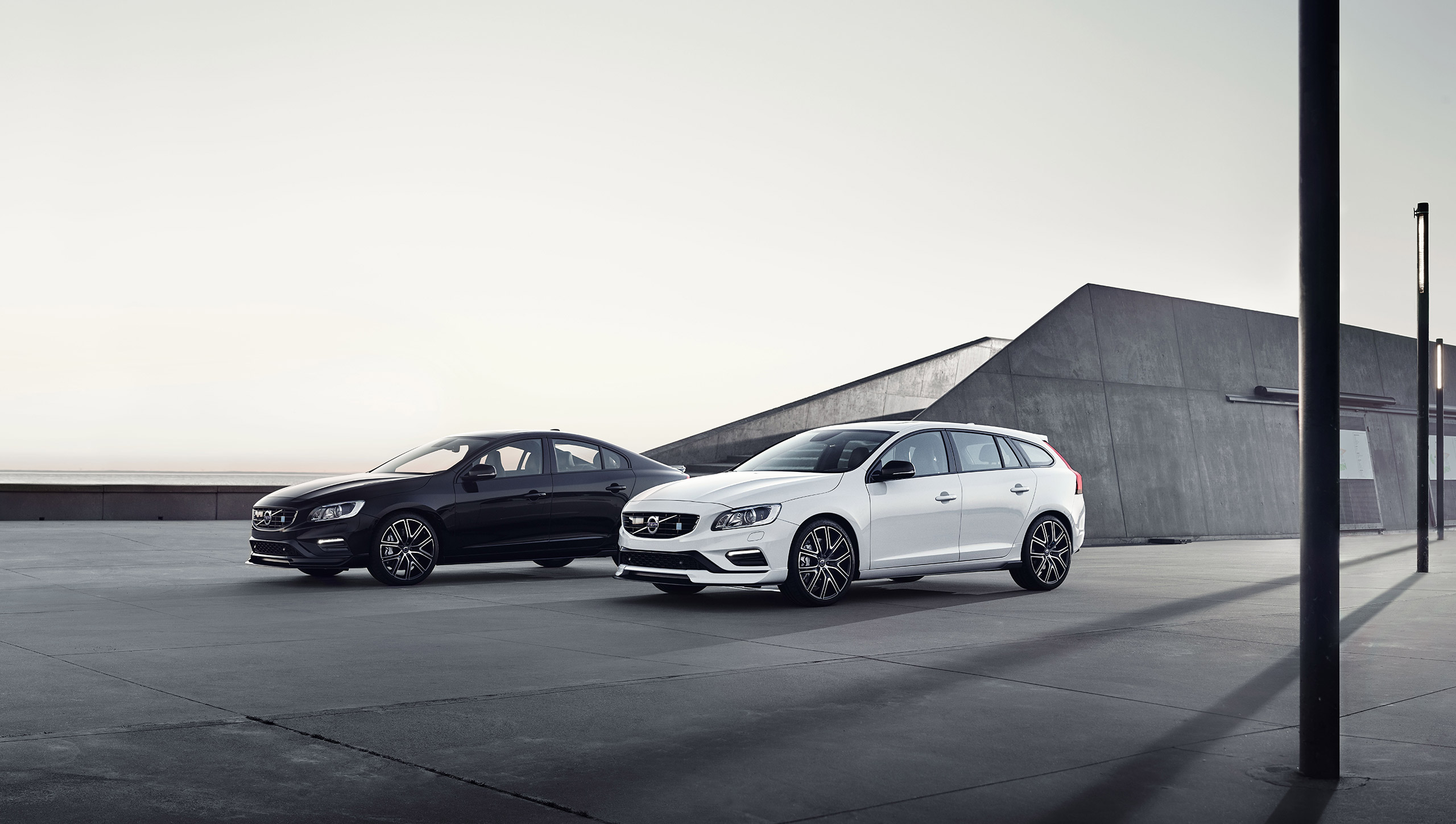 Musta Volvo S60 ja valkoinen Volvo V60 parkissa tasanteella, takana geometrinen betonirakennus.