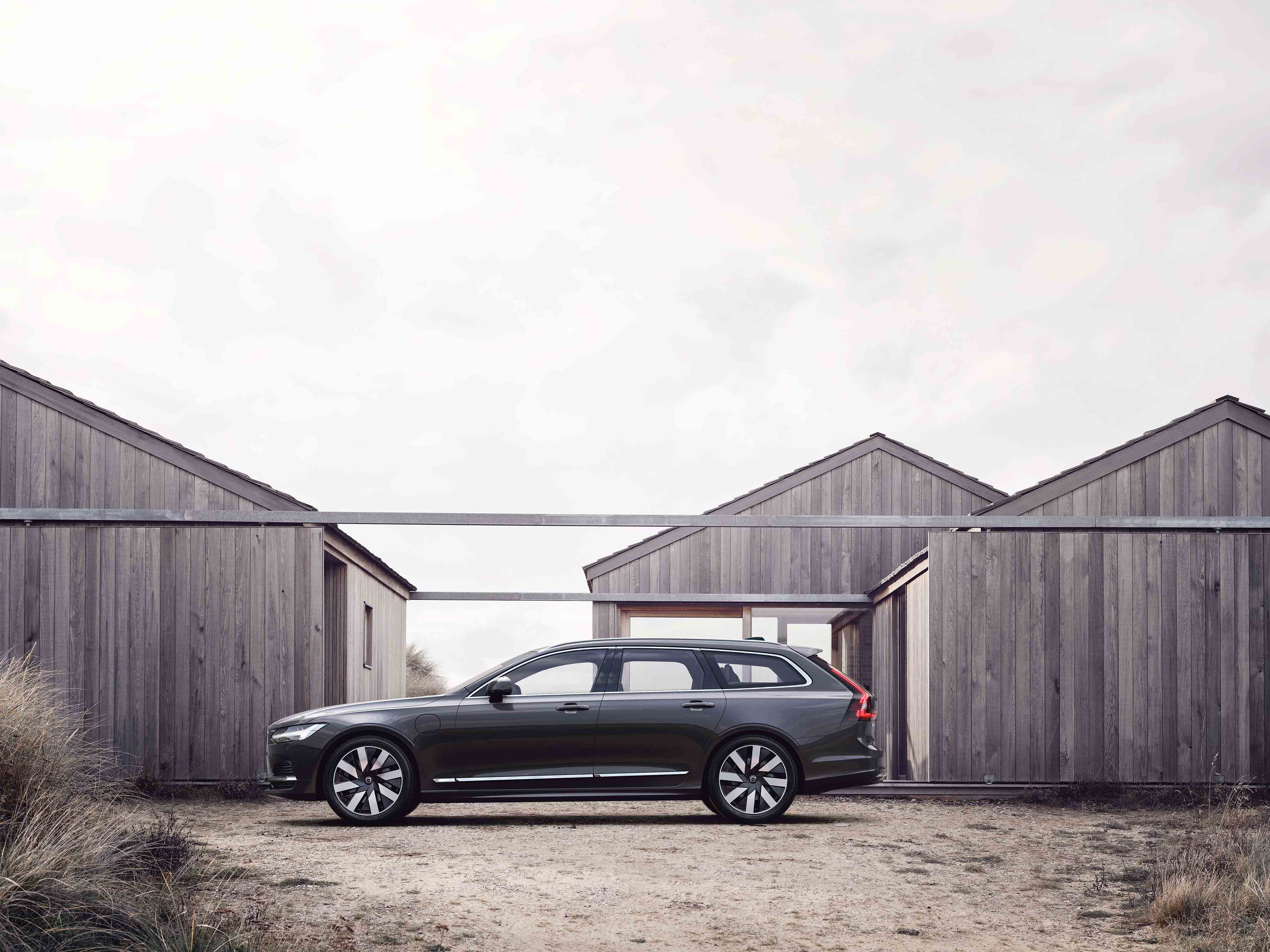 Tumman harma Volvo V90 parkkeerattuna harmaiden lautatalojen eteen.