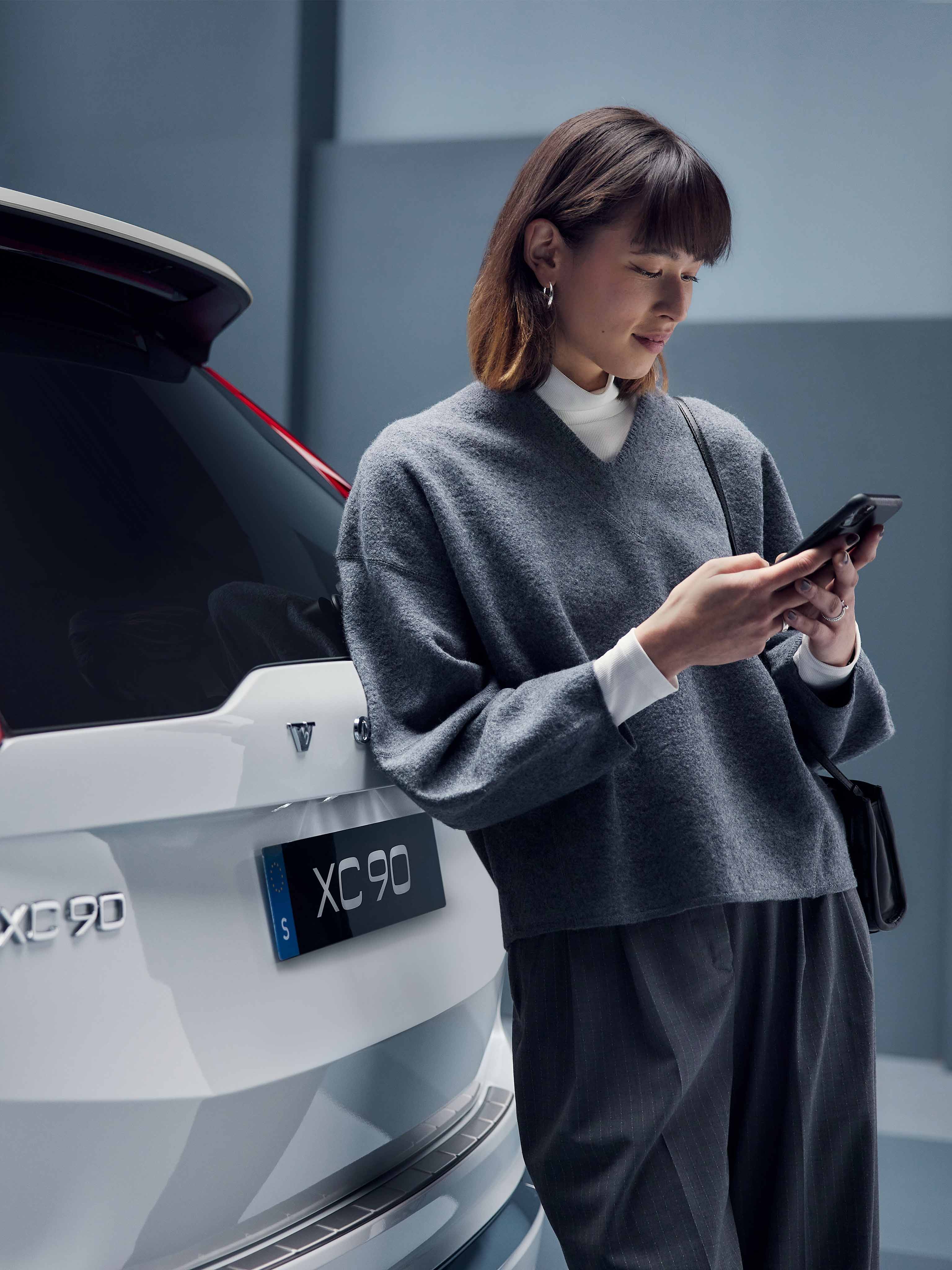 Nainen nojaa valkean Volvo XC90:en takaluukkuun ja selaa puhelimella Volvo Cars sovellusta.