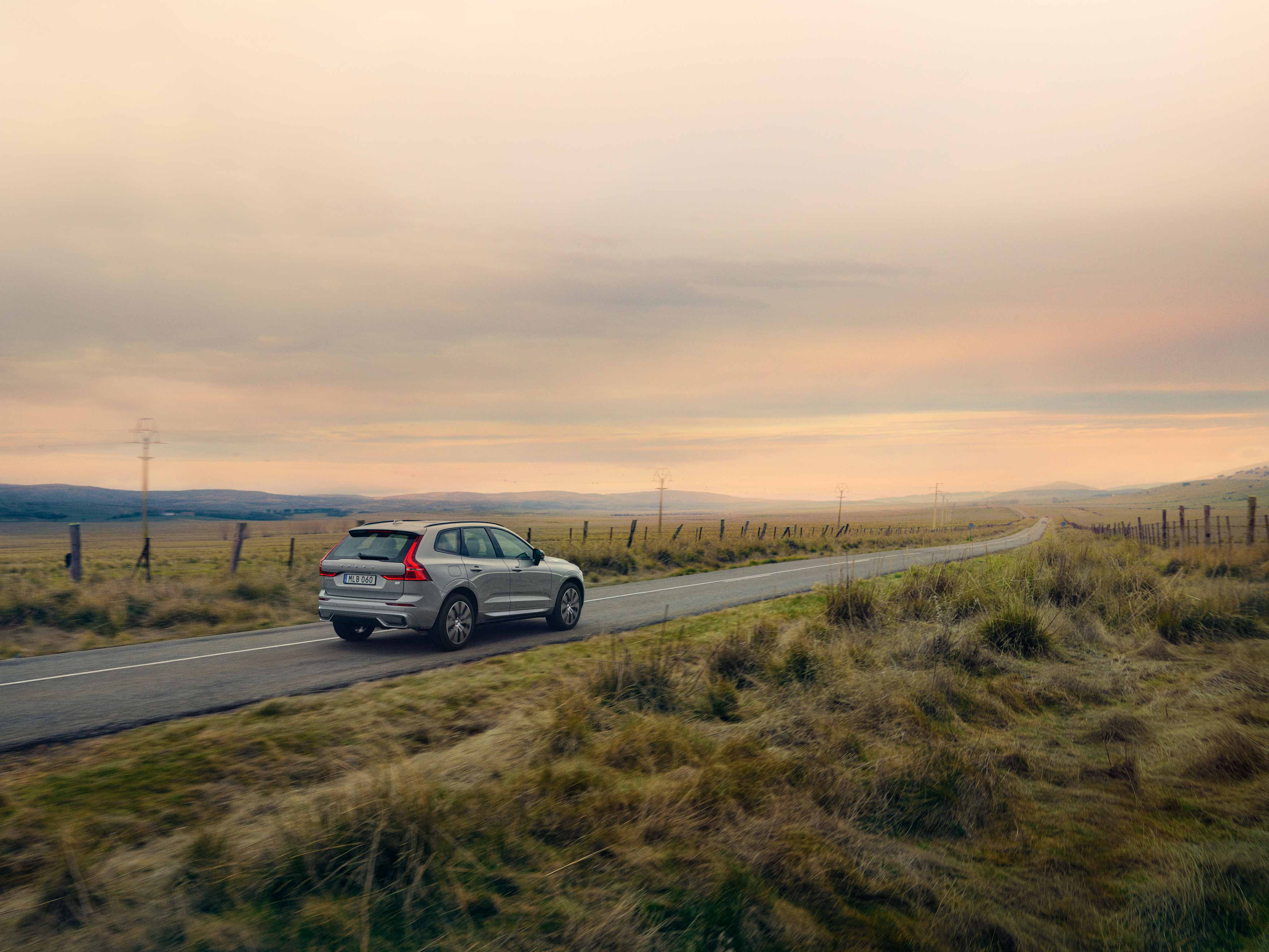 Silver color Volvo XC60 Recharge driving between fields towards sunset