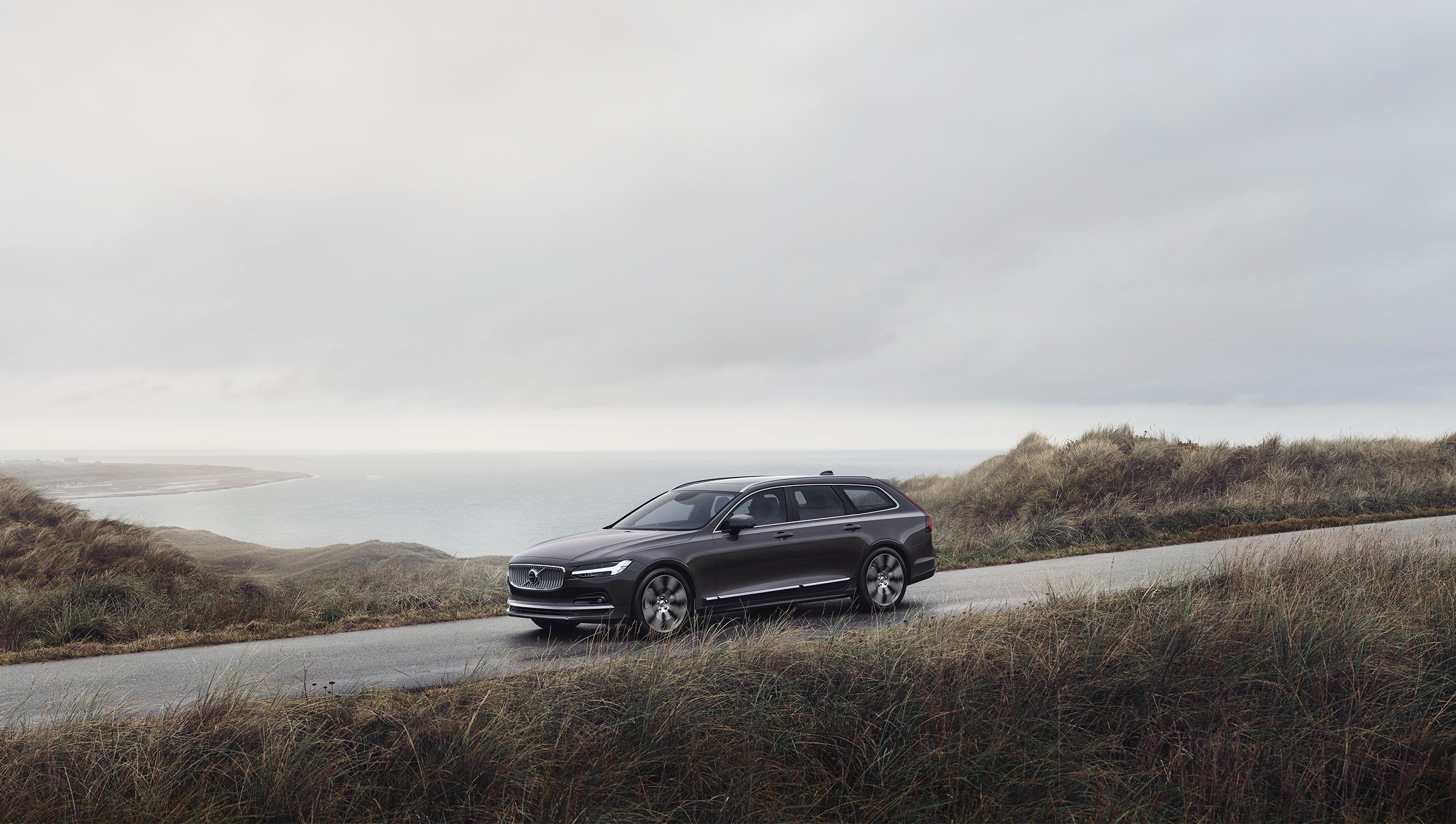 Platinum grey Volvo V90 driving on mountains with seaview.