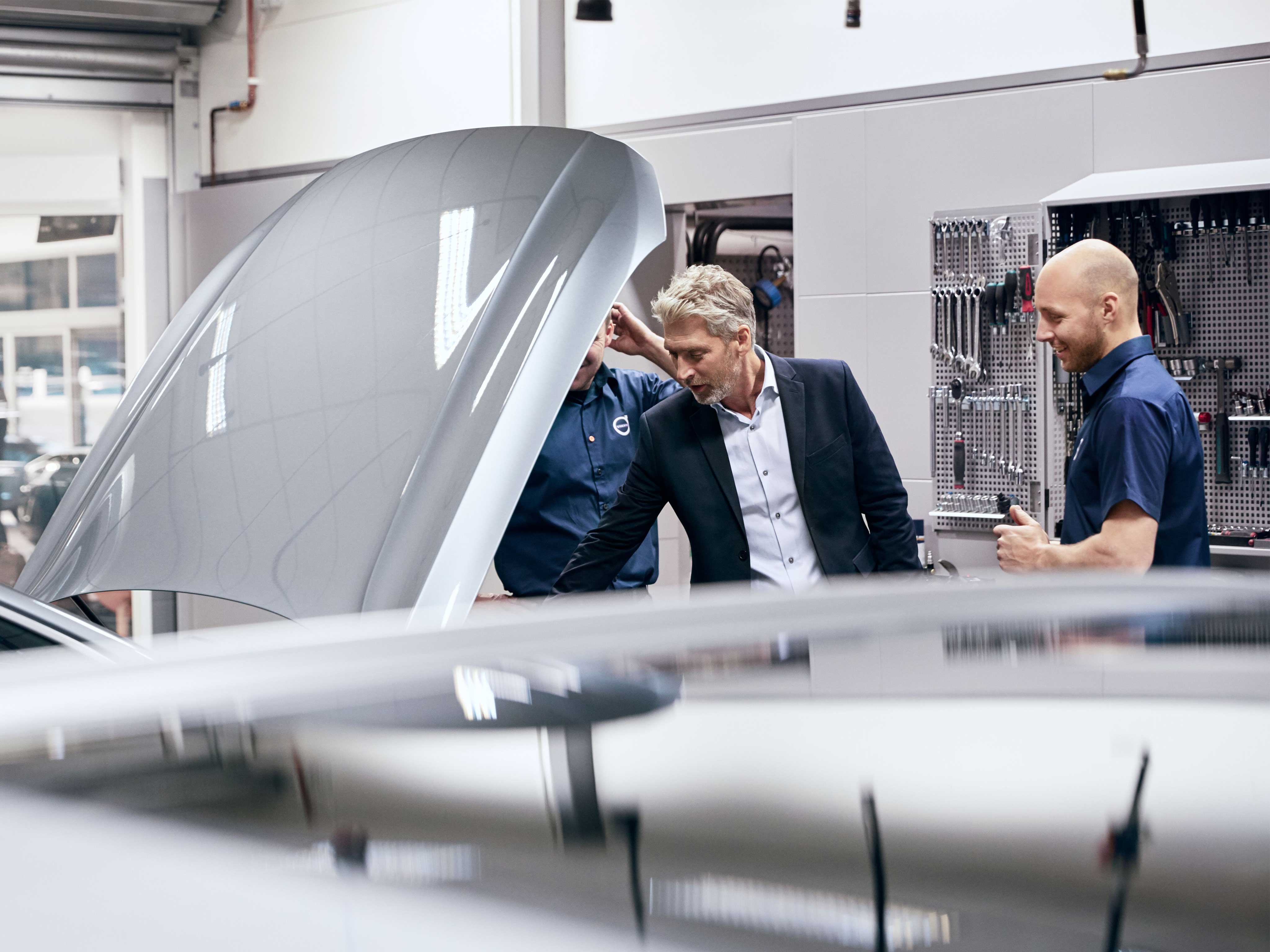 Two Volvo mechanics and a man looking at a car with open hood.