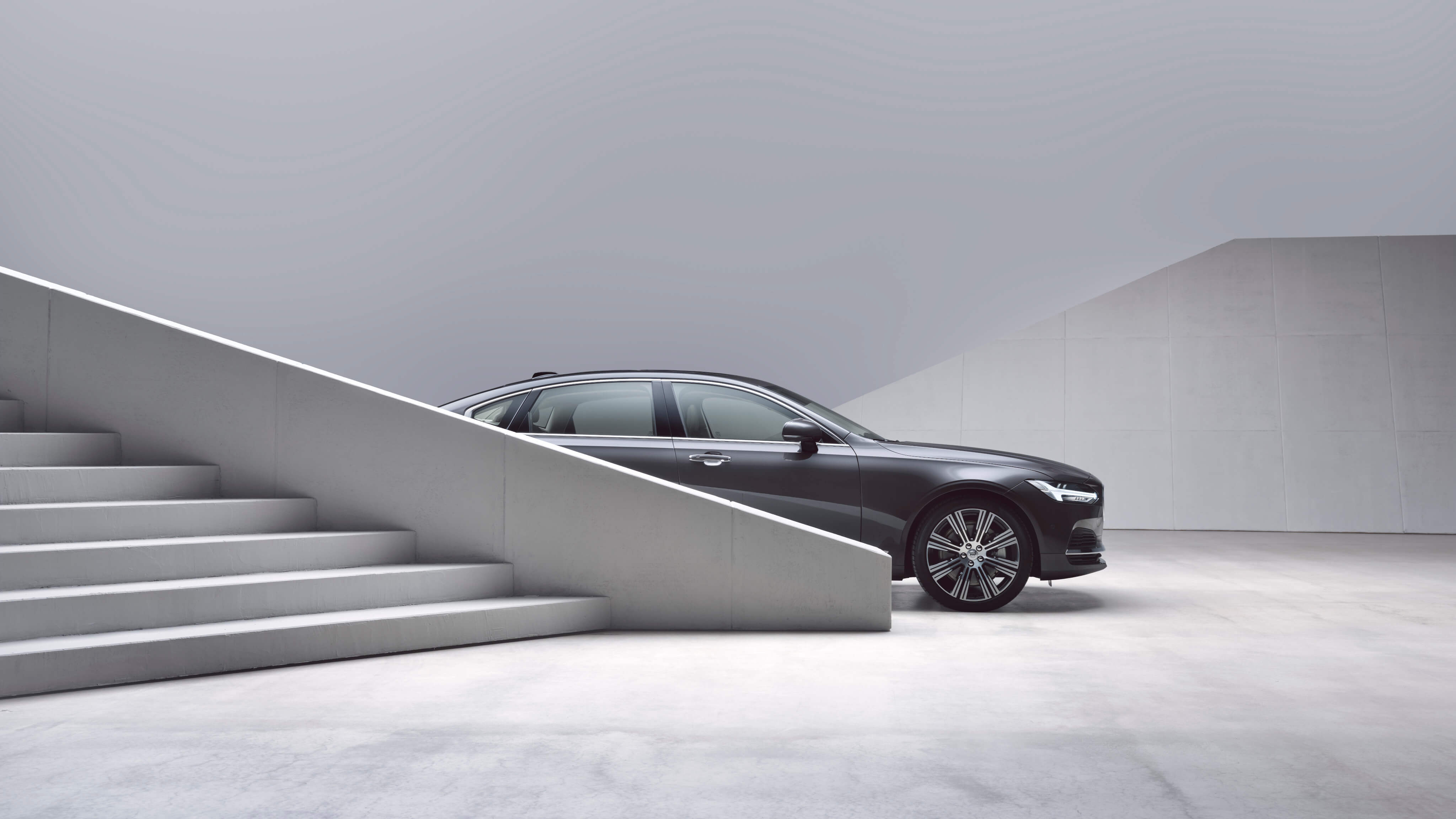 Volvo S90 Recharge Inscription Platinum Grey steht in einer grauen Umgebung hinter Treppen - Seitenschuss