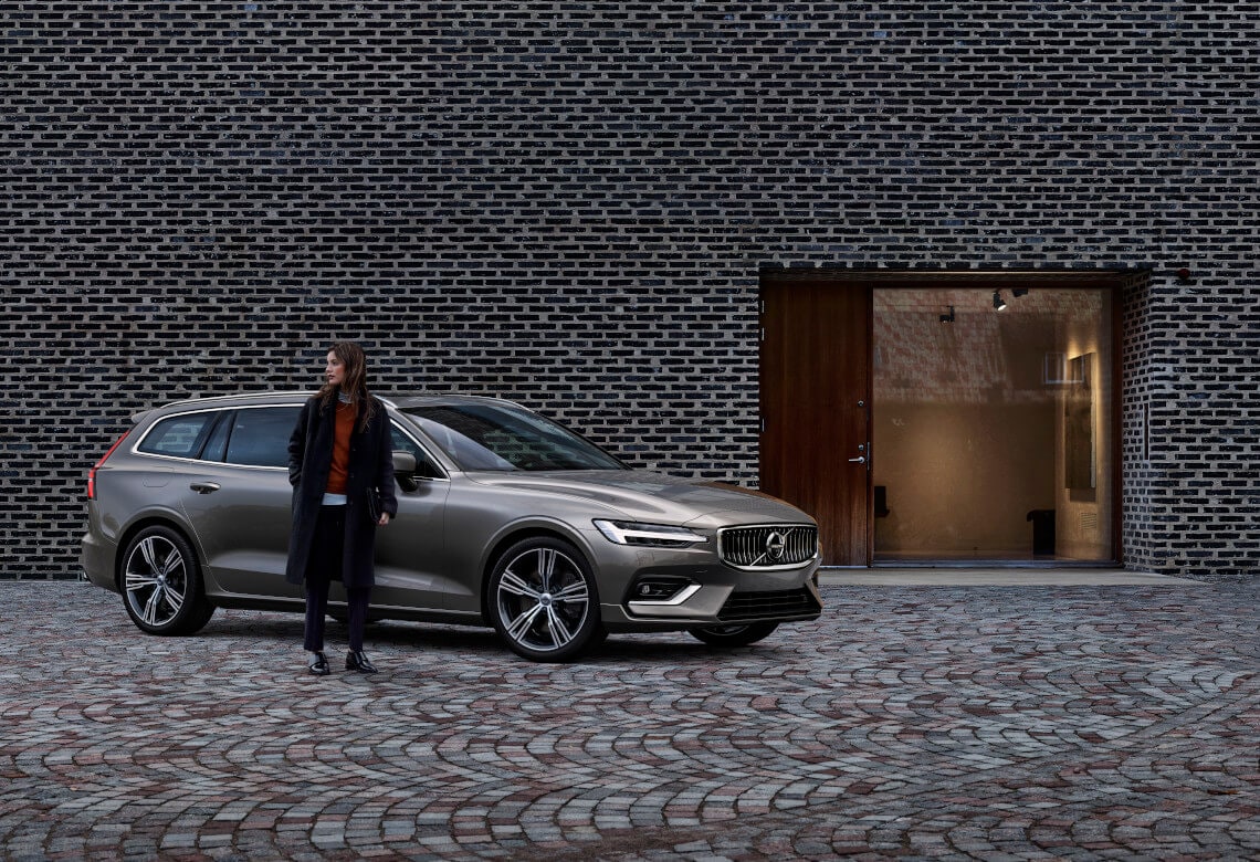 Frau steht vor einem Volvo V60 Inscription in Pebble Grey in einer Hauseinfahrt