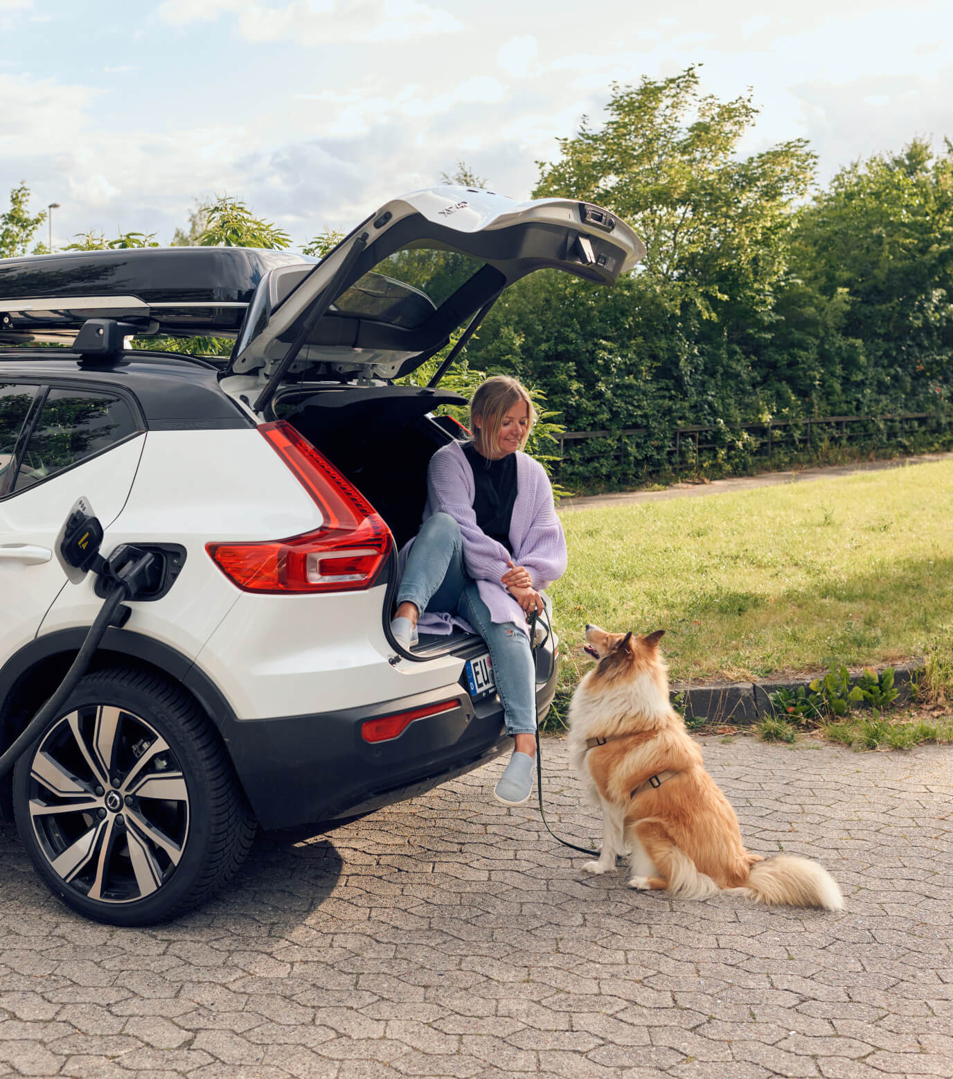 Eine Frau wartet im Kofferraum ihres vollelektrischen Volvo, bis dieser an der Ladesäule aufgeladen ist. Vor dem Fahrzeug sitzt ein Hund.