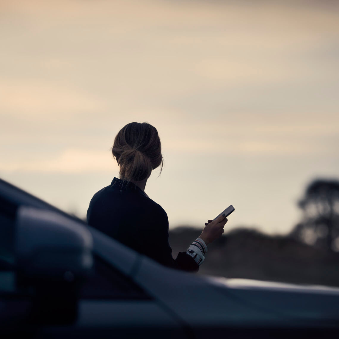Frau steht bei Sonnenuntergang mit ihrem Handy an einem Volvo