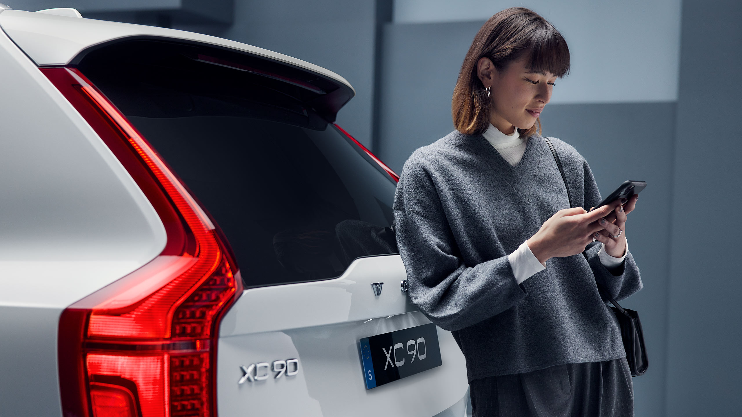 Jeune femme regardant son smartphone à coté d’une Volvo XC90 blanche.