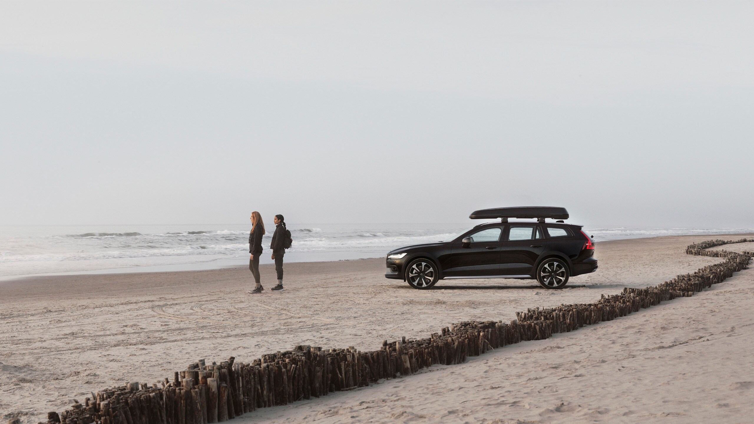 Dos chicas en la playa con un Volvo V90 y un cofre de techo