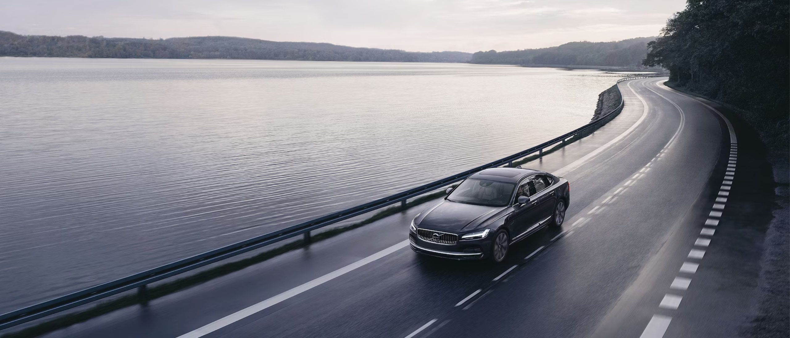 Una Volvo S90 nera sta percorrendo una strada che costeggia un grande lago.